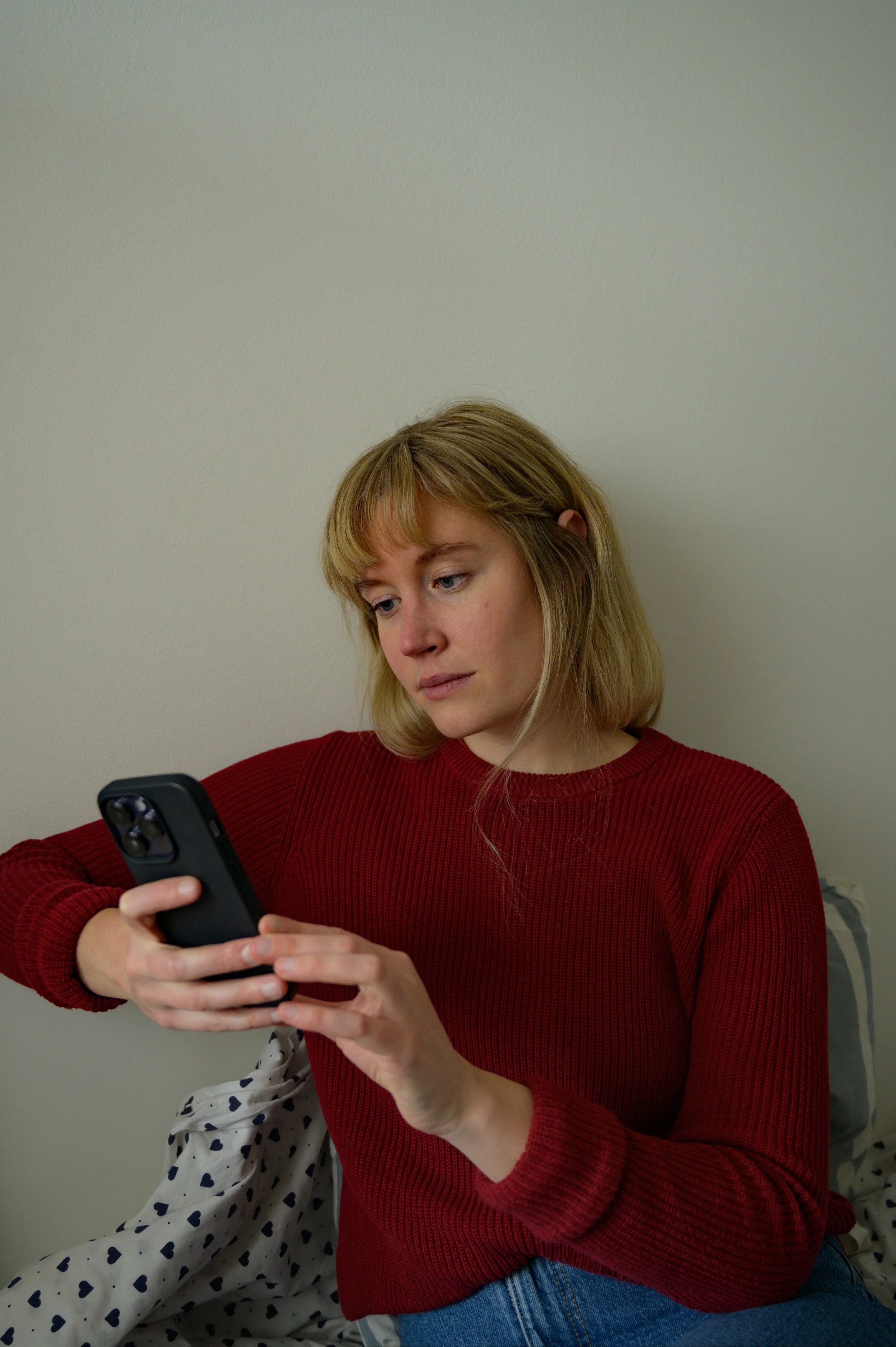 A woman with blonde hair and bangs is pictured from the waist up, sitting against a white wall. She is looking intently at the screen of a smartphone held in both hands. She wears a red knit sweater and blue jeans, with a heart-patterned pillow visib