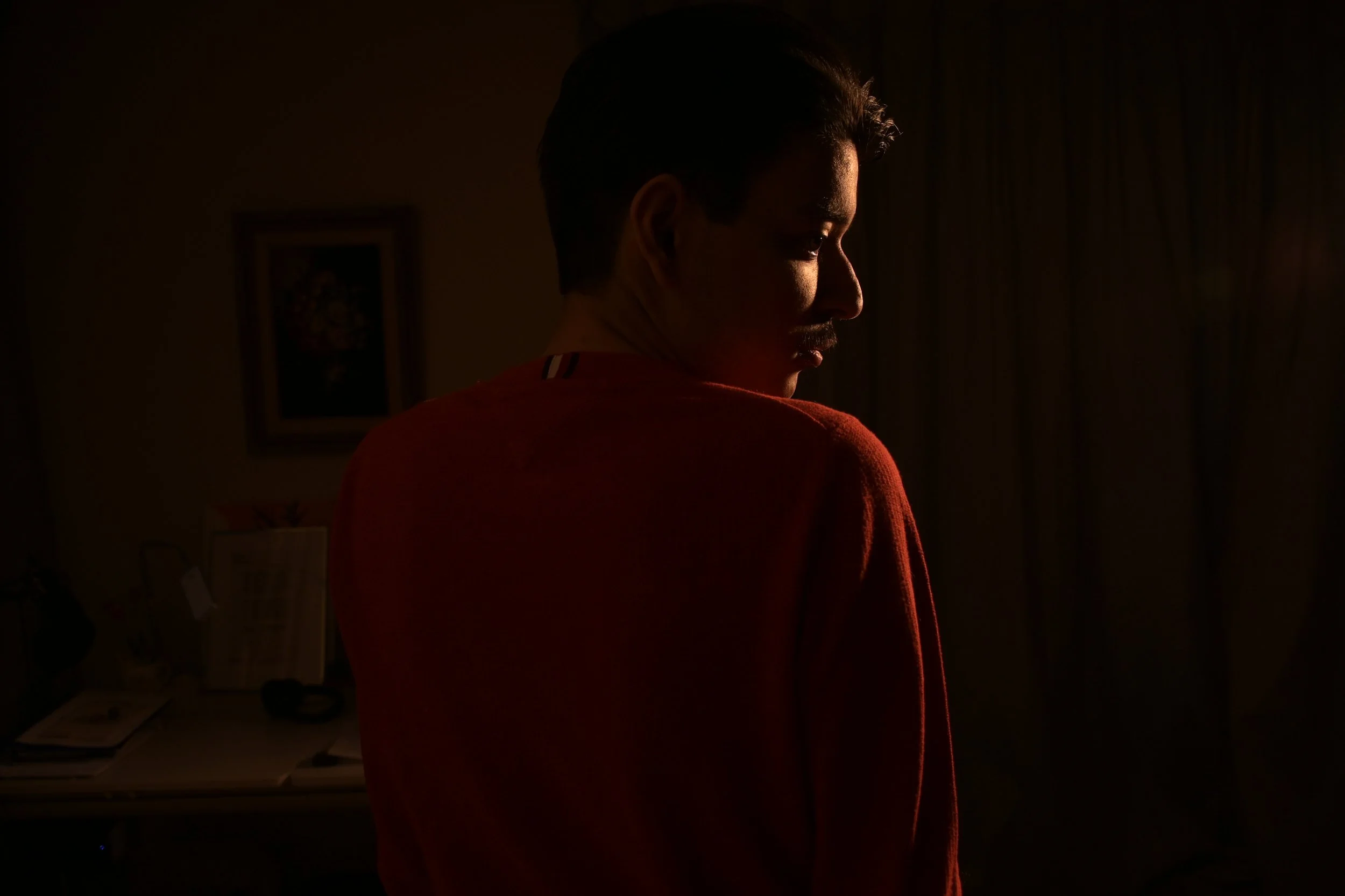 A young man in a red shirt standing in a room with dim lighting, facing sideways with a serious expression.