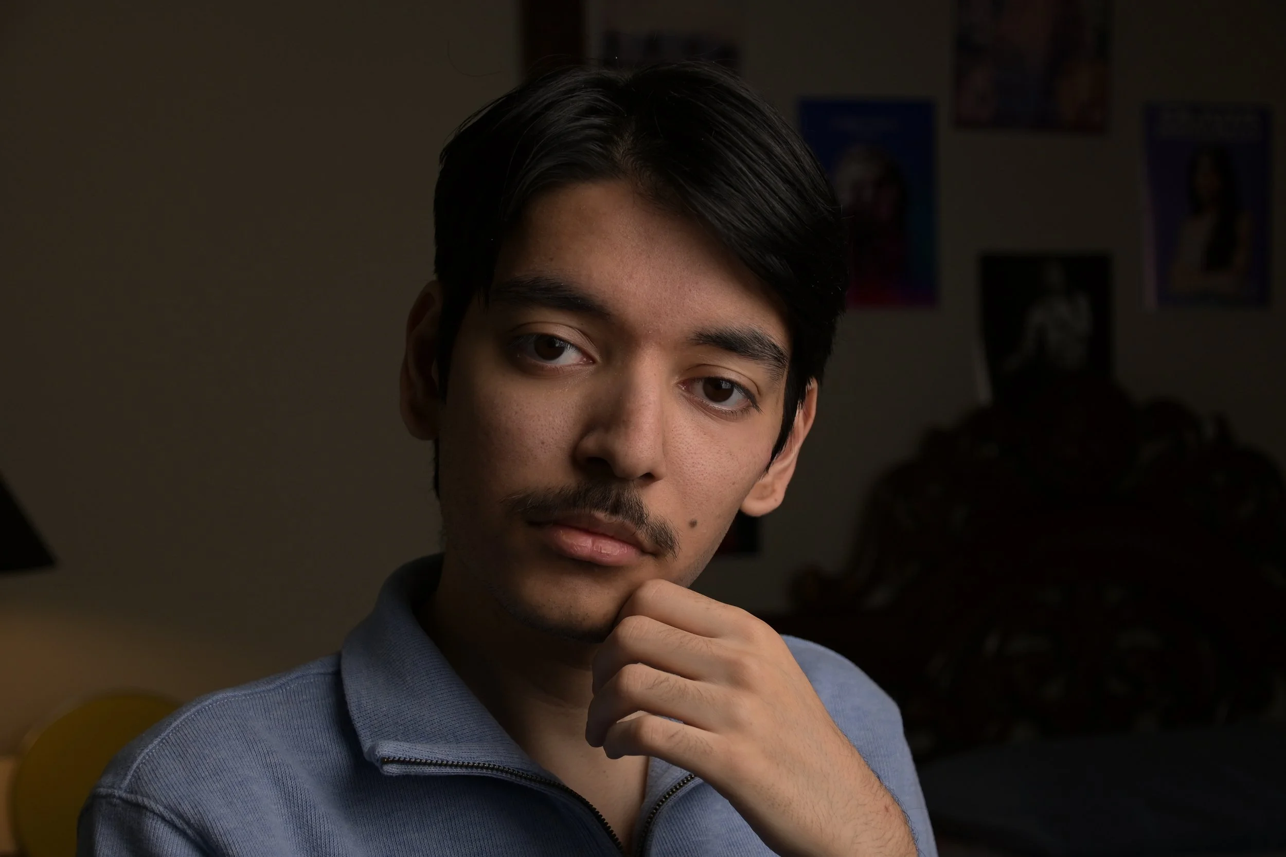 A young man with black hair and a mustache, wearing a blue zip-up jacket, looking thoughtfully at the camera with his hand resting on his chin.
