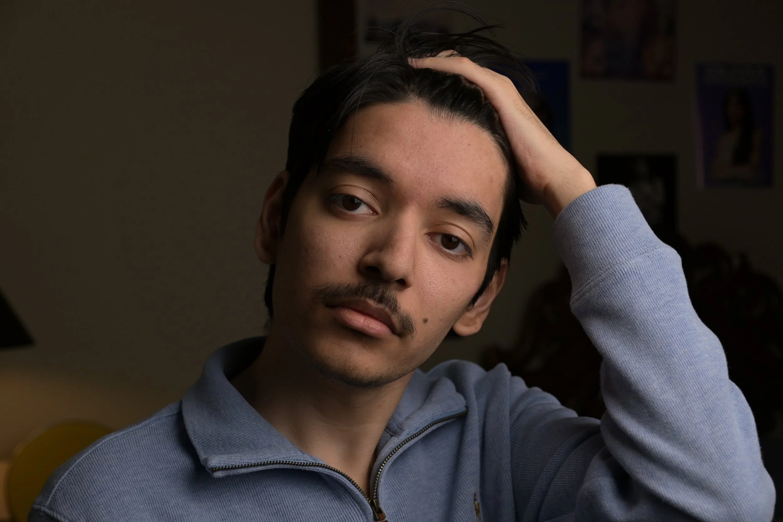 Young man with dark hair and light skin, holding his head with one hand, wearing a gray zip-up jacket, in an indoor setting.