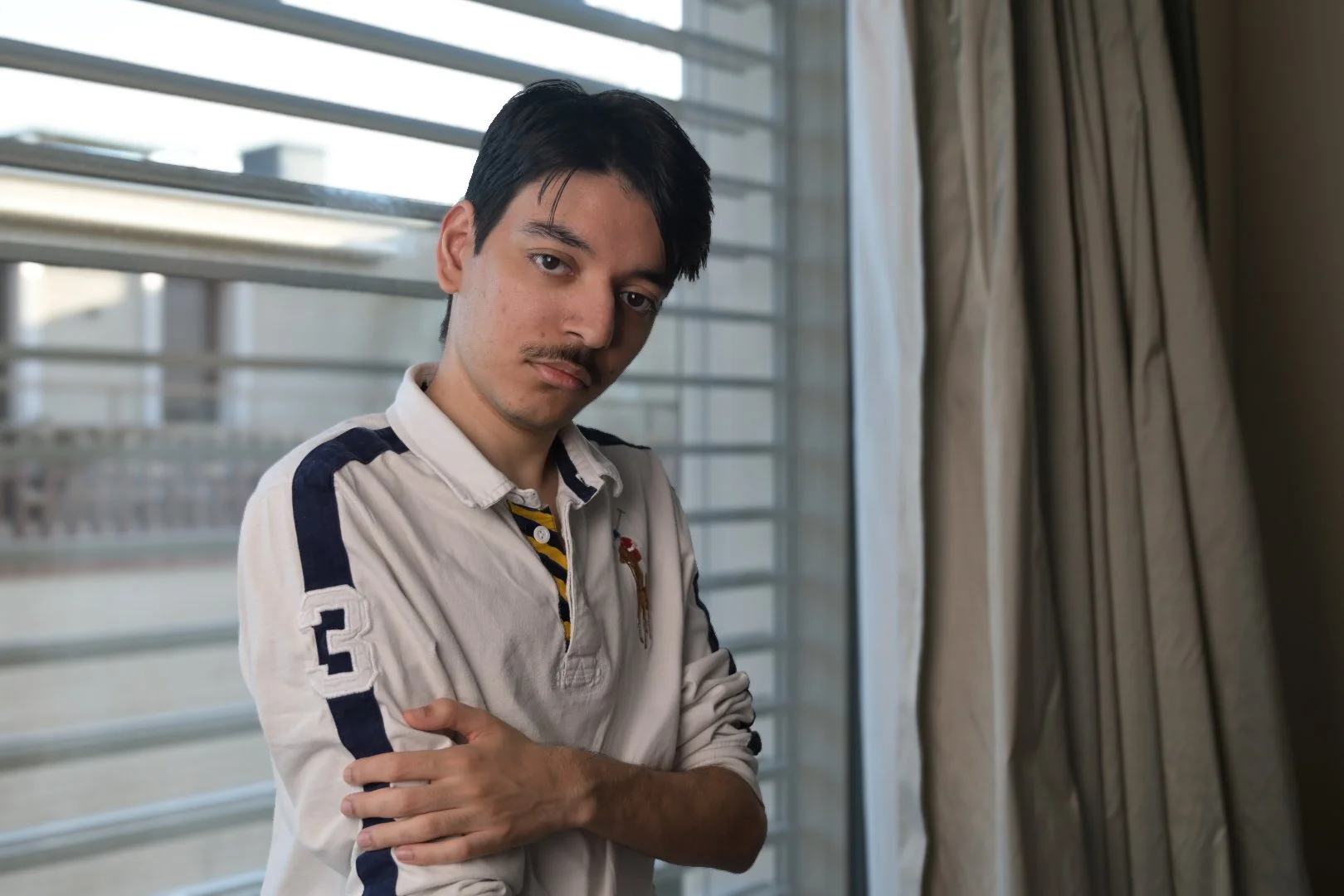 A young man with dark hair and a mustache, looking at the camera with a serious expression, standing indoors by a window with closed blinds and curtains.