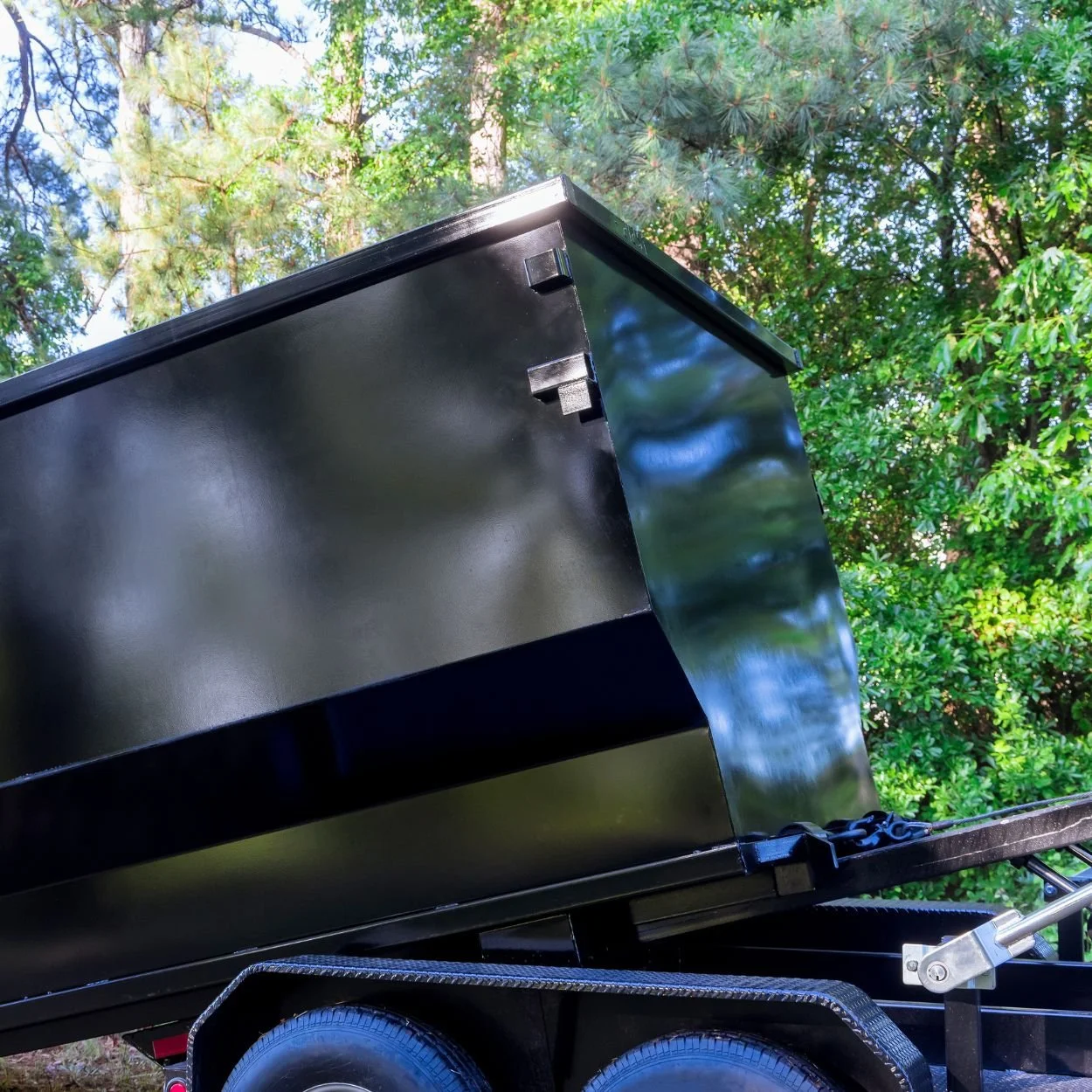 A black metal trailer with a large black box on top, set outdoors among green trees and foliage.