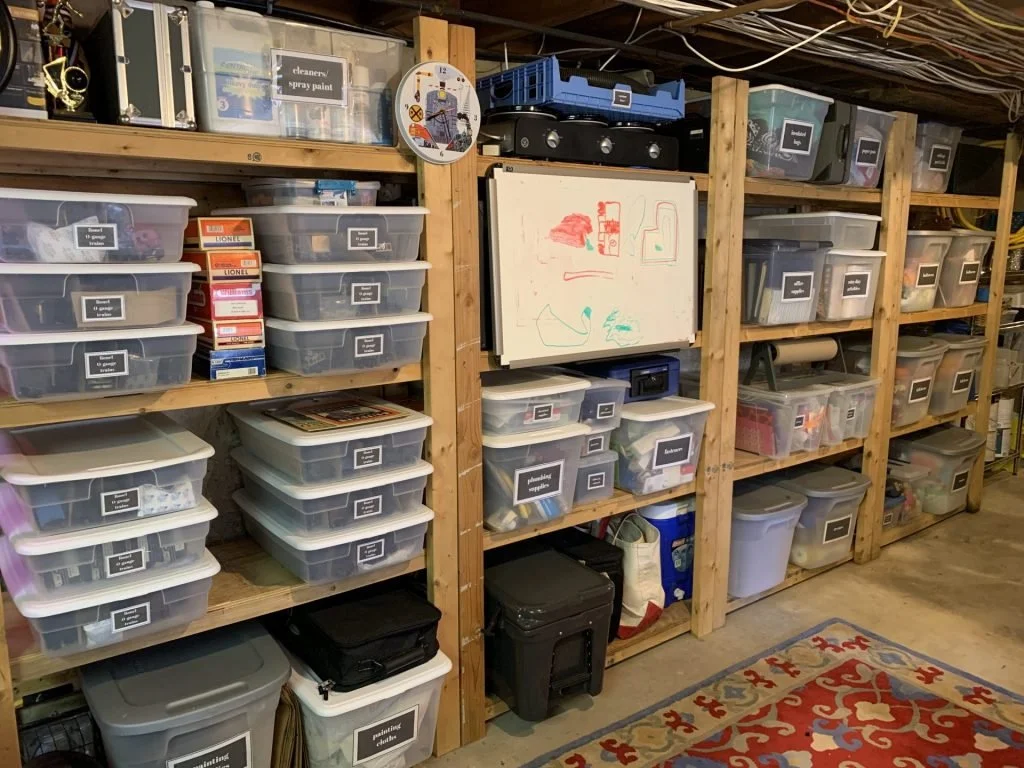Clear storage bins stacked on wooden basement shelves with visible labels for easy identification.