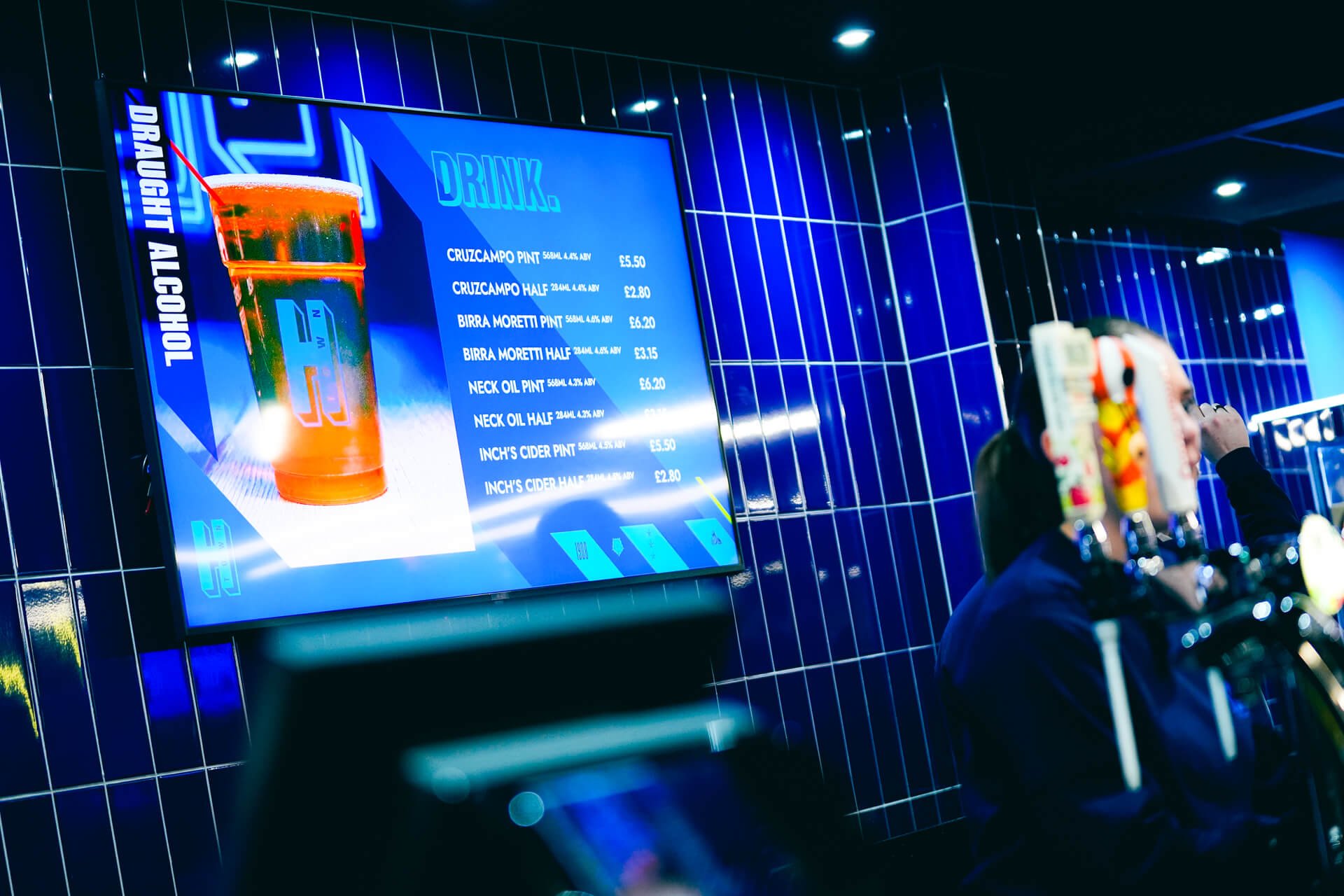 A digital menu board displaying various beers and prices at a bar, with a blue tiled wall in the background and drink taps in the foreground.