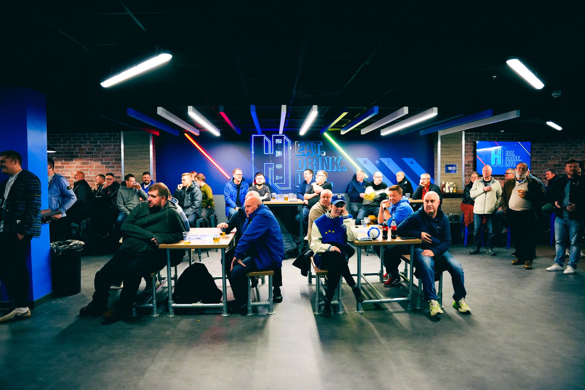 People sitting and standing in a sports bar with neon signs that say "Eat. Drink. Live." and a large screen displaying a football game.