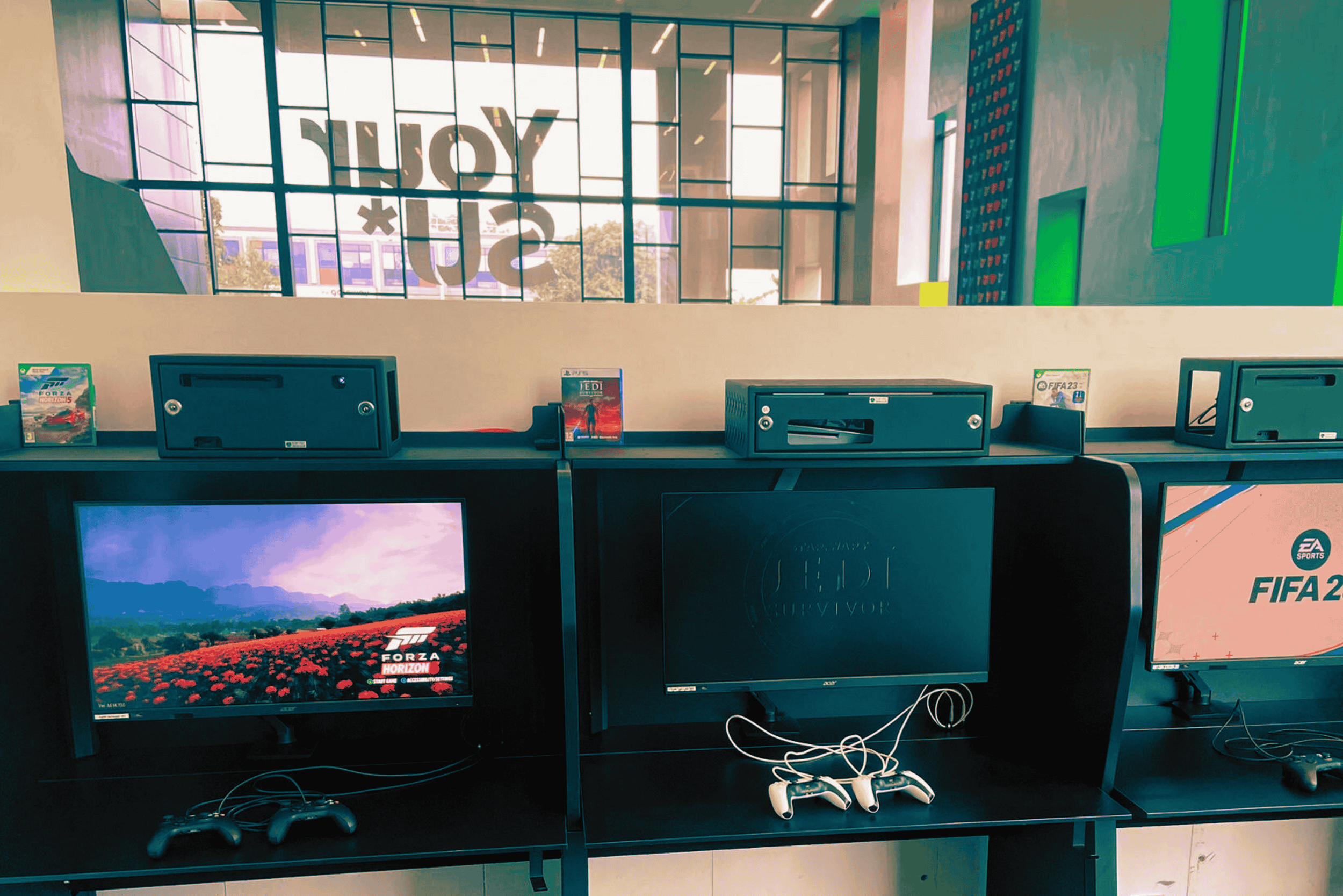 Video game station with two screens, controllers, and stacks of video game cases, with a large glass window in the background showing the words 'TOY STORY'.