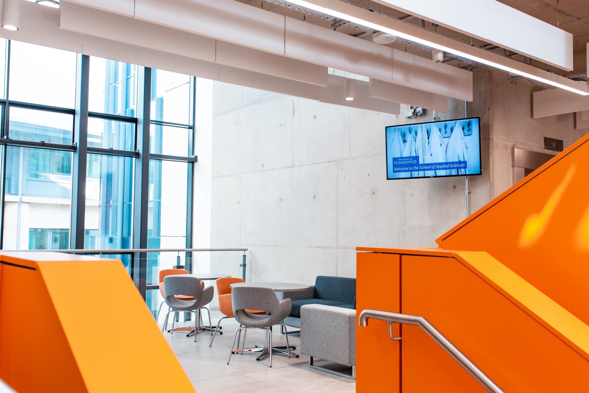 Modern lounge area with orange stairs, gray and orange chairs, round tables, large windows, and a digital screen displaying a welcome message at the University of Huddersfield School of Applied Sciences.
