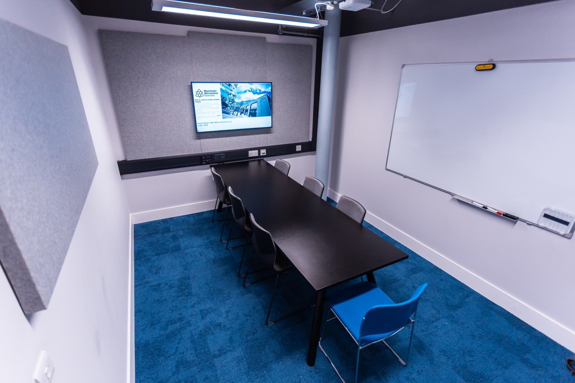 Small conference room with a black table, six black chairs, one blue chair, a television screen displaying a presentation, a whiteboard on the wall, and gray soundproofing panels, blue carpeted floor.