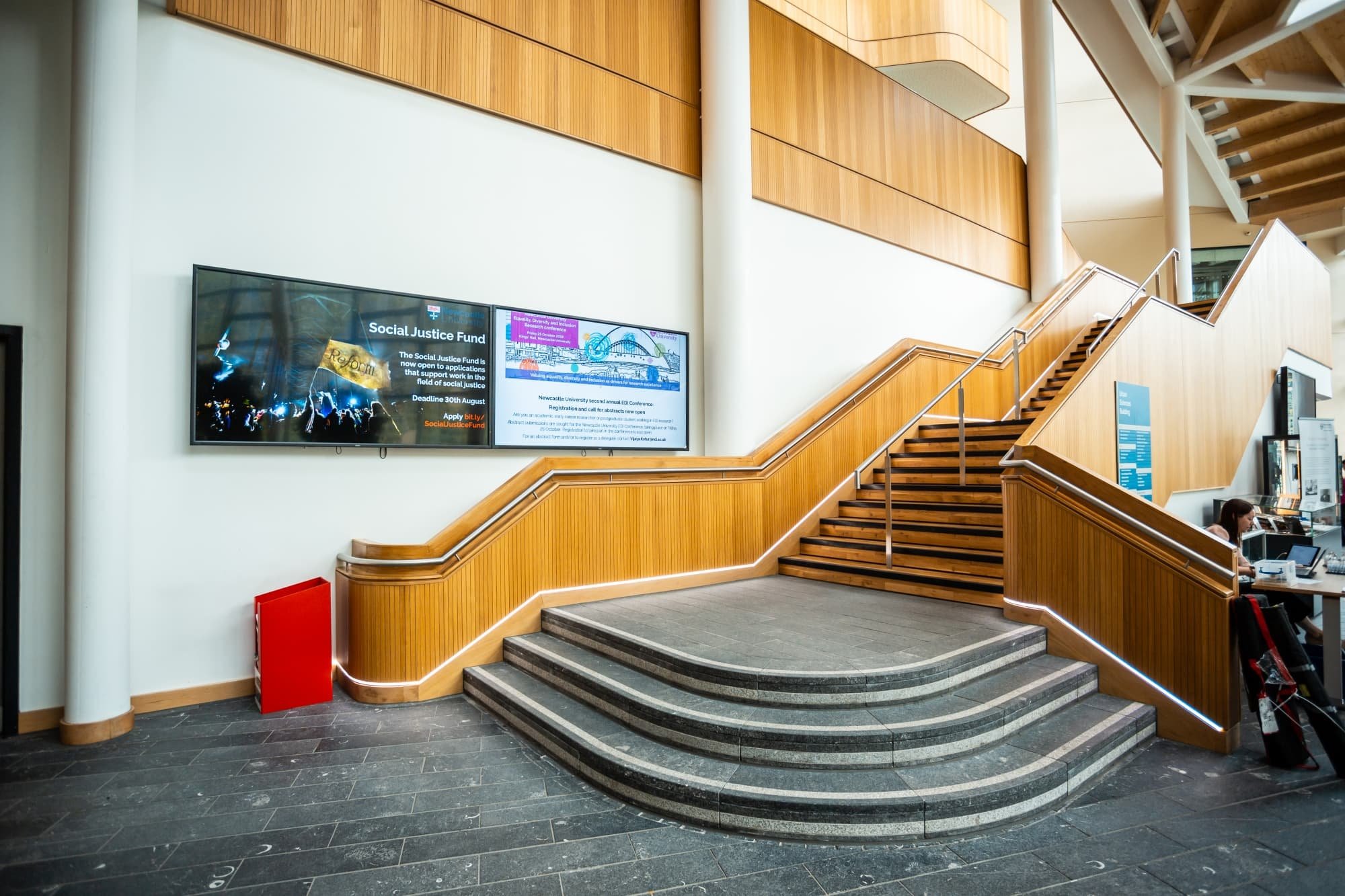 Interior of a modern building with wooden stairs, a digital display on the wall, and a person working at a desk.
