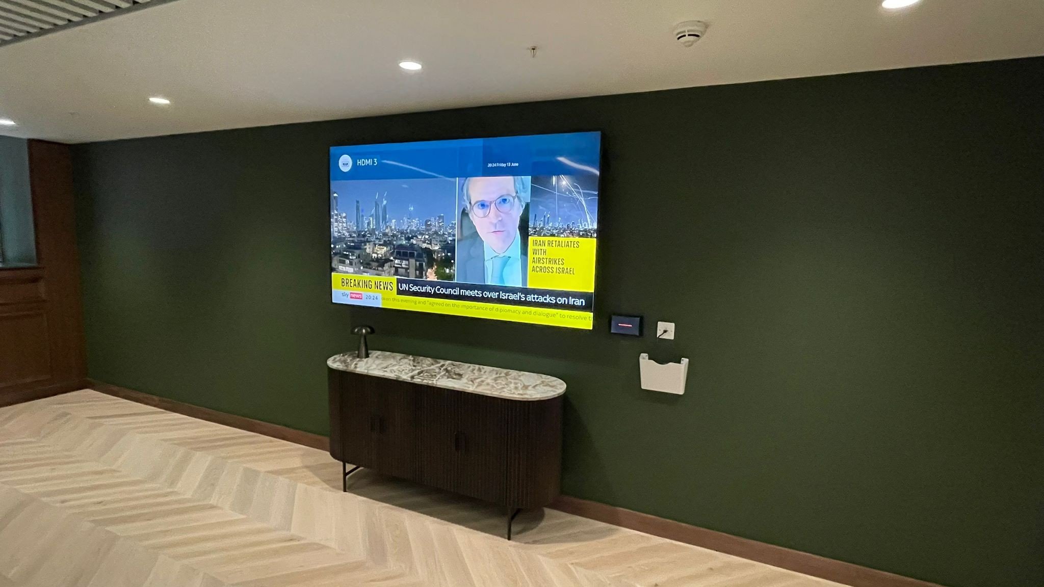 Modern living room with a large wall-mounted flat-screen TV displaying a news broadcast, a wooden sideboard with a marble top underneath the TV, and wall outlets and a white box mounted on the green wall. The room has light-colored wood flooring and