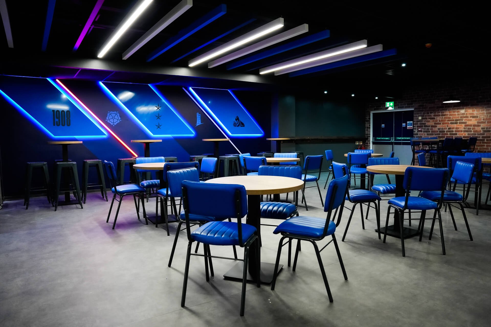 An empty bar or lounge area with blue chairs and tables, decorated with colorful neon lights on a dark wall, and a brick wall on the right side.