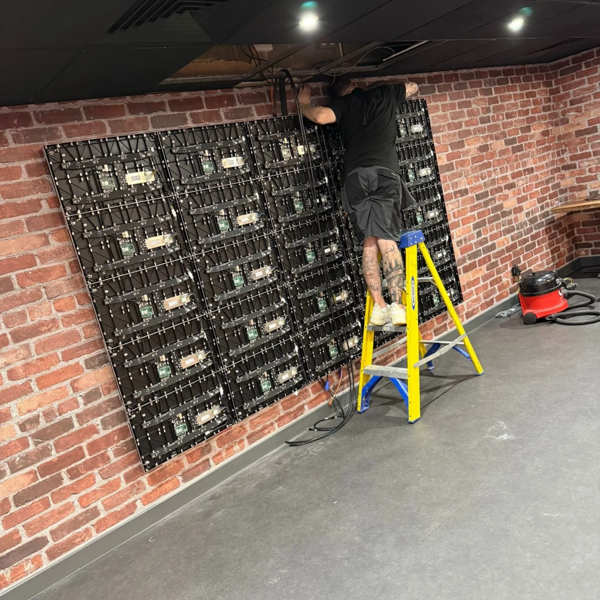 A person working on a large LED display mounted on a brick wall, standing on a yellow step ladder, with a vacuum cleaner nearby.