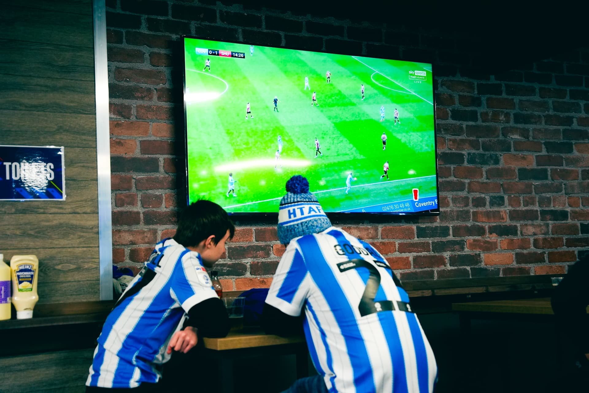 Two young men wearing blue and white striped football jerseys and a beanie watching a soccer game on a flat-screen TV mounted on a brick wall in a bar or sports lounge.