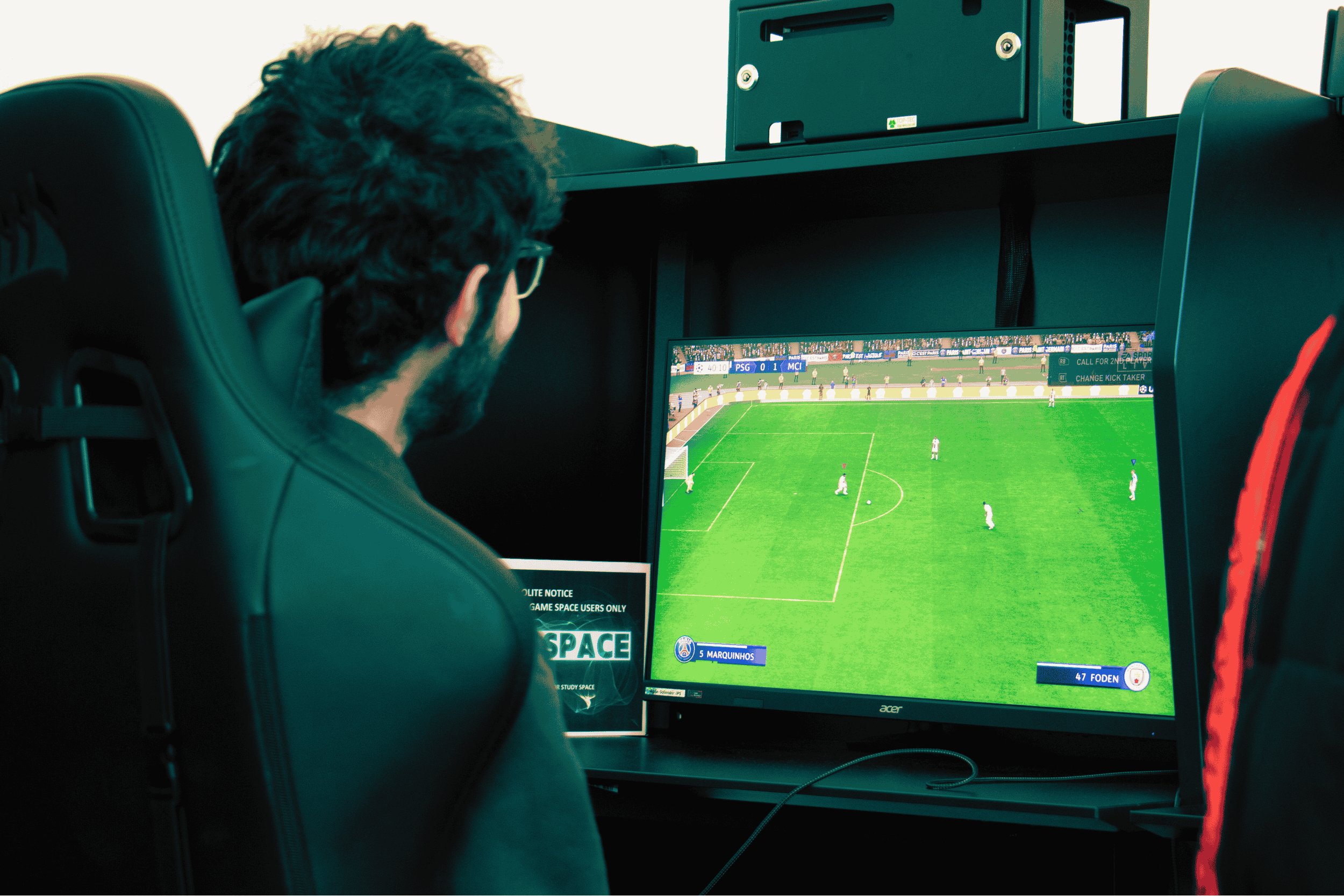 A man with dark hair and glasses playing a soccer video game on a computer monitor, sitting in a gaming chair.