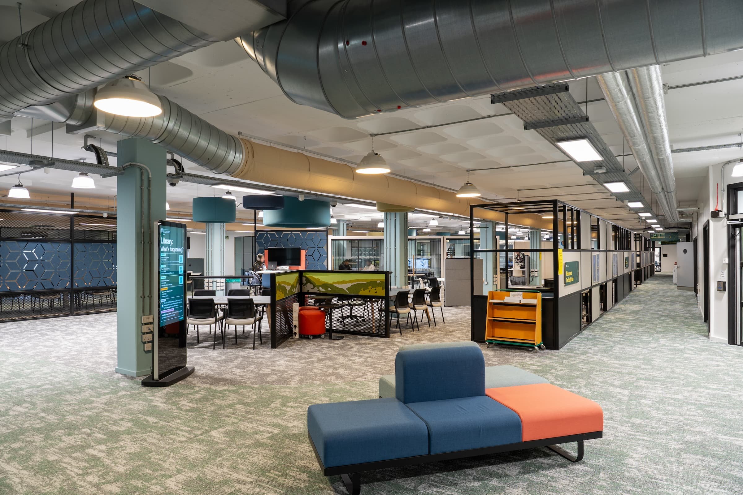 Modern library or office lounge with a blue and orange couch in the foreground, tables with chairs, a digital directory, and partitioned study areas in the background, featuring exposed ceiling ducts and industrial lighting.
