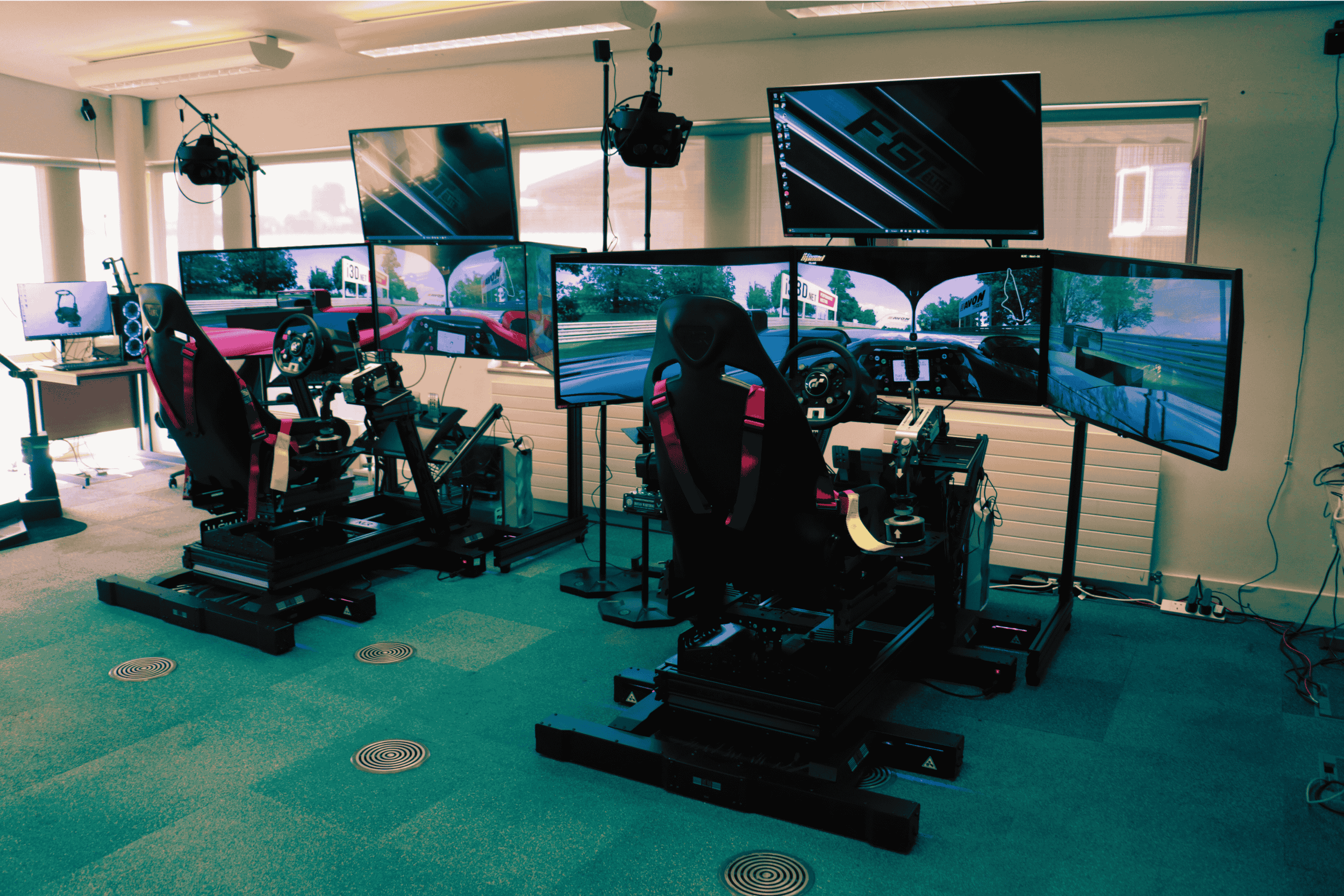 A racing simulator with two racing seats, each equipped with multiple large monitors displaying a racing game, force feedback steering wheels, pedals, and shifters. The setup includes microphones, speakers, and a computer on a desk to the side.