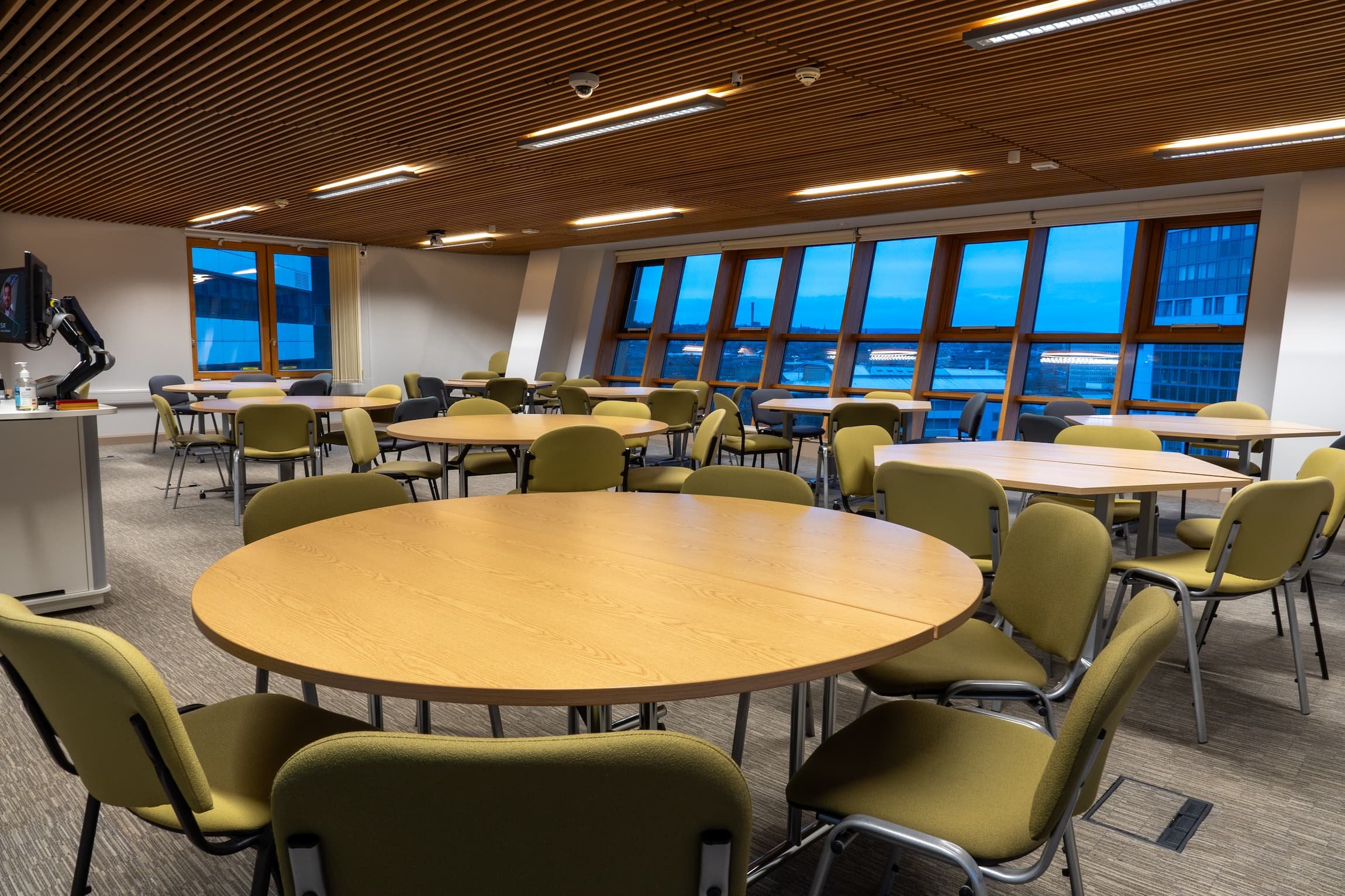 Empty conference or meeting room with round and rectangular tables, green and gray chairs, large windows with city view, wooden ceiling with lighting.