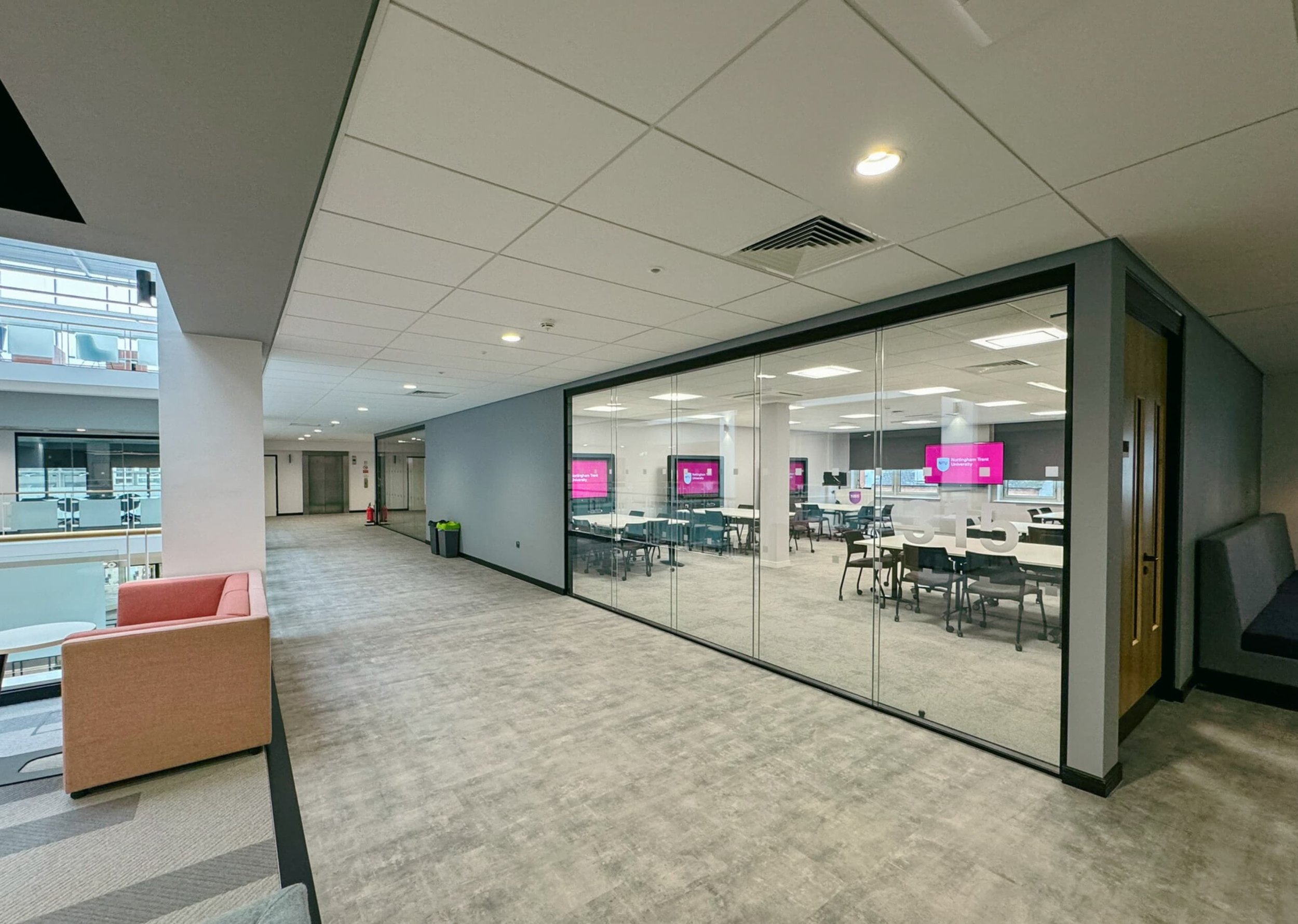 Interior view of an office corridor with a glass-walled conference room, modern chairs, and digital screens, illuminated by ceiling lights.