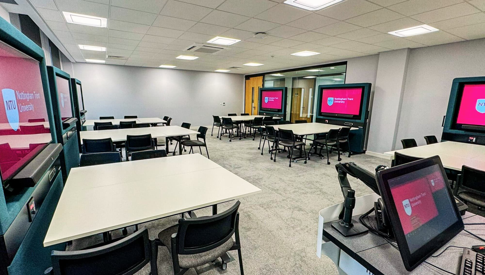 Empty classroom with multiple white tables, black chairs, and four large television screens displaying the Nottingham Trent University logo in red and purple, in a modern educational space.