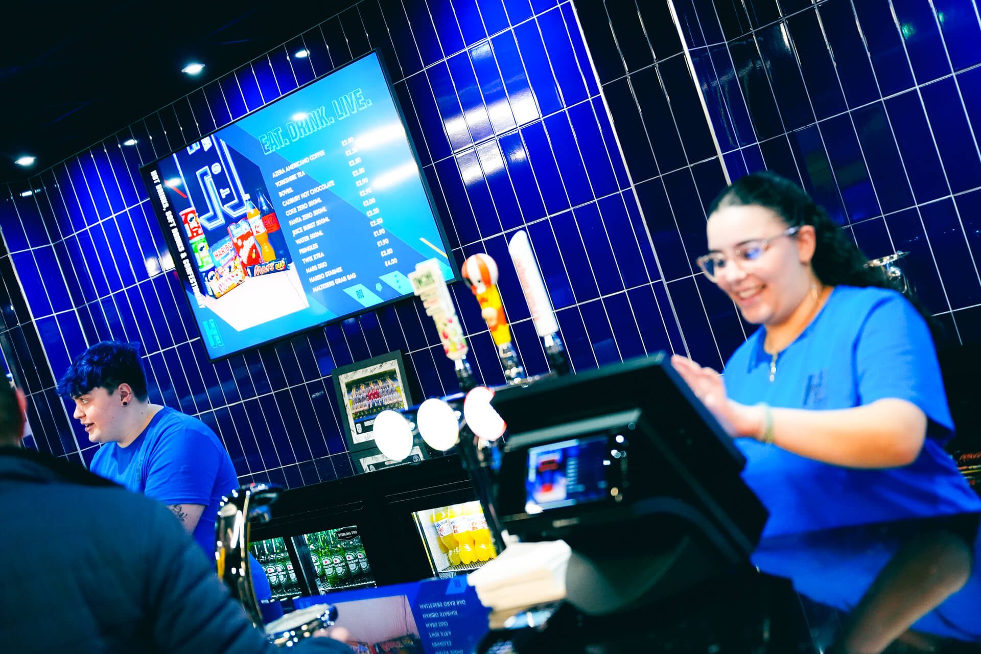 A woman smiling behind a cash register at a bar counter in a lively, modern establishment with a blue tiled wall and a digital menu screen.
