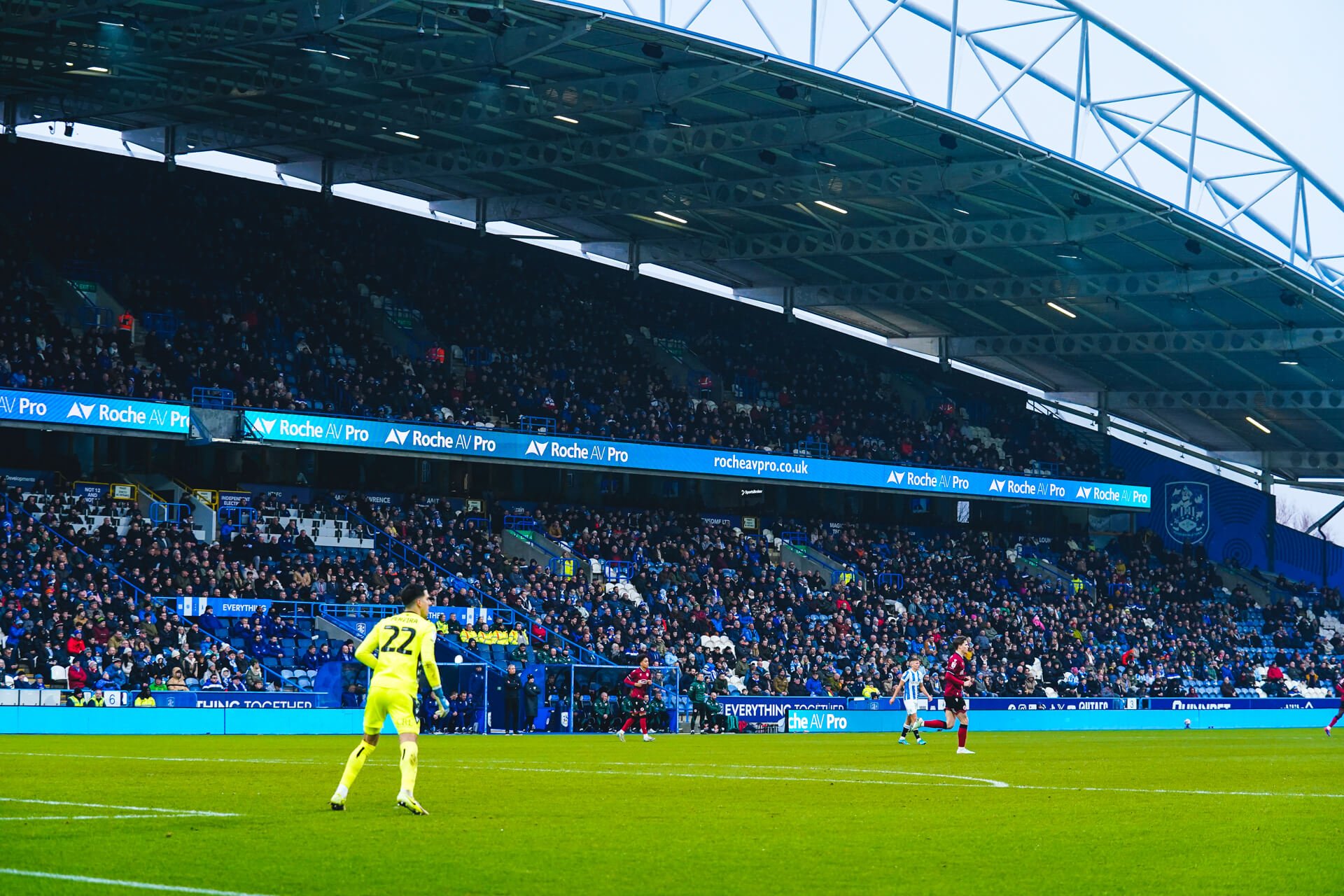 A soccer match in progress at a large stadium with a crowded audience. Players are on the field, with one goalkeeper in yellow, and others in red and blue uniforms. The stadium has digital advertising banners and a modern roof structure.
