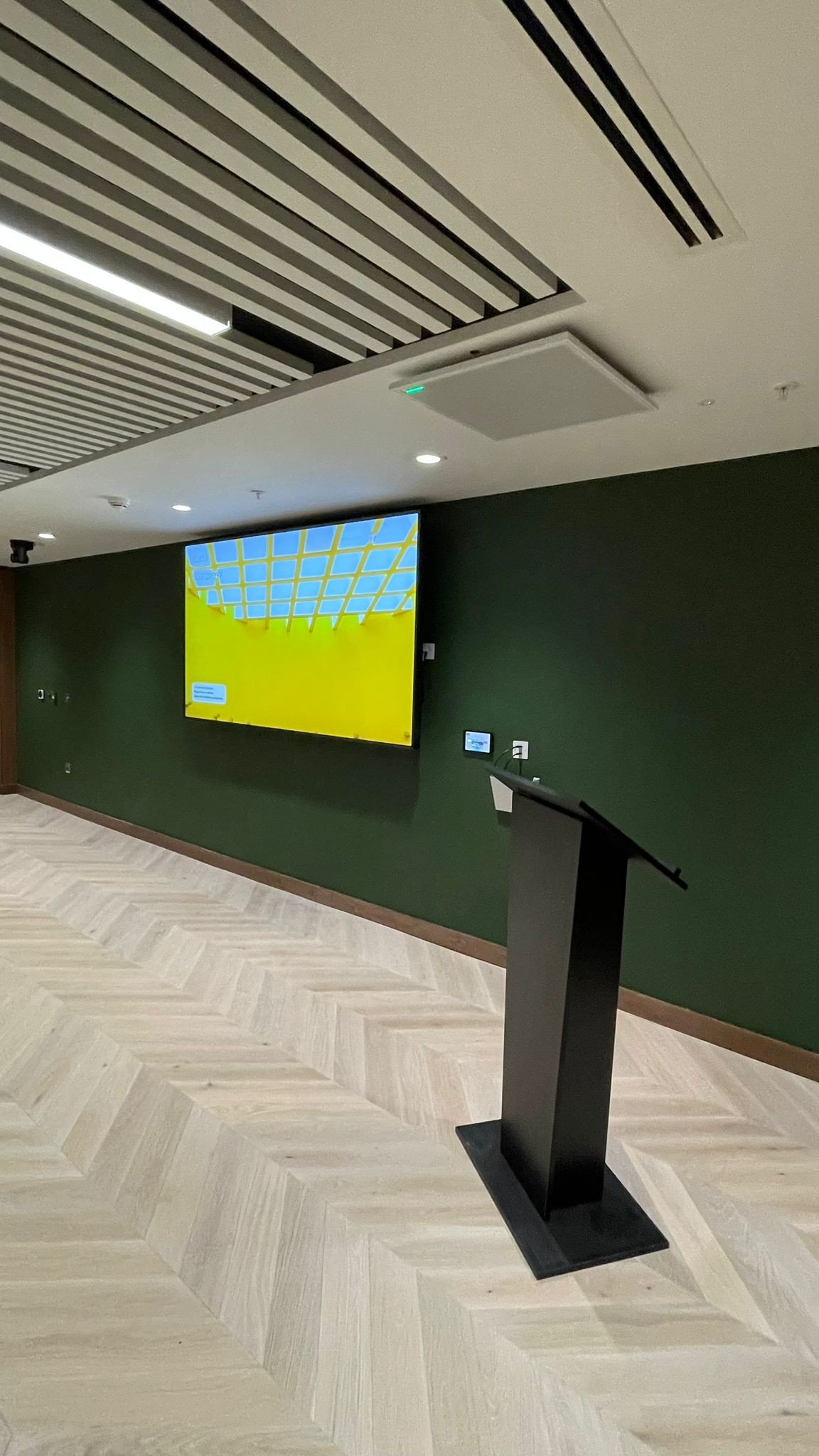 Empty presentation room with black podium, large screen displaying yellow and blue grid pattern, dark green wall, light wood herringbone floor, and ceiling with linear lighting and ventilation panels.