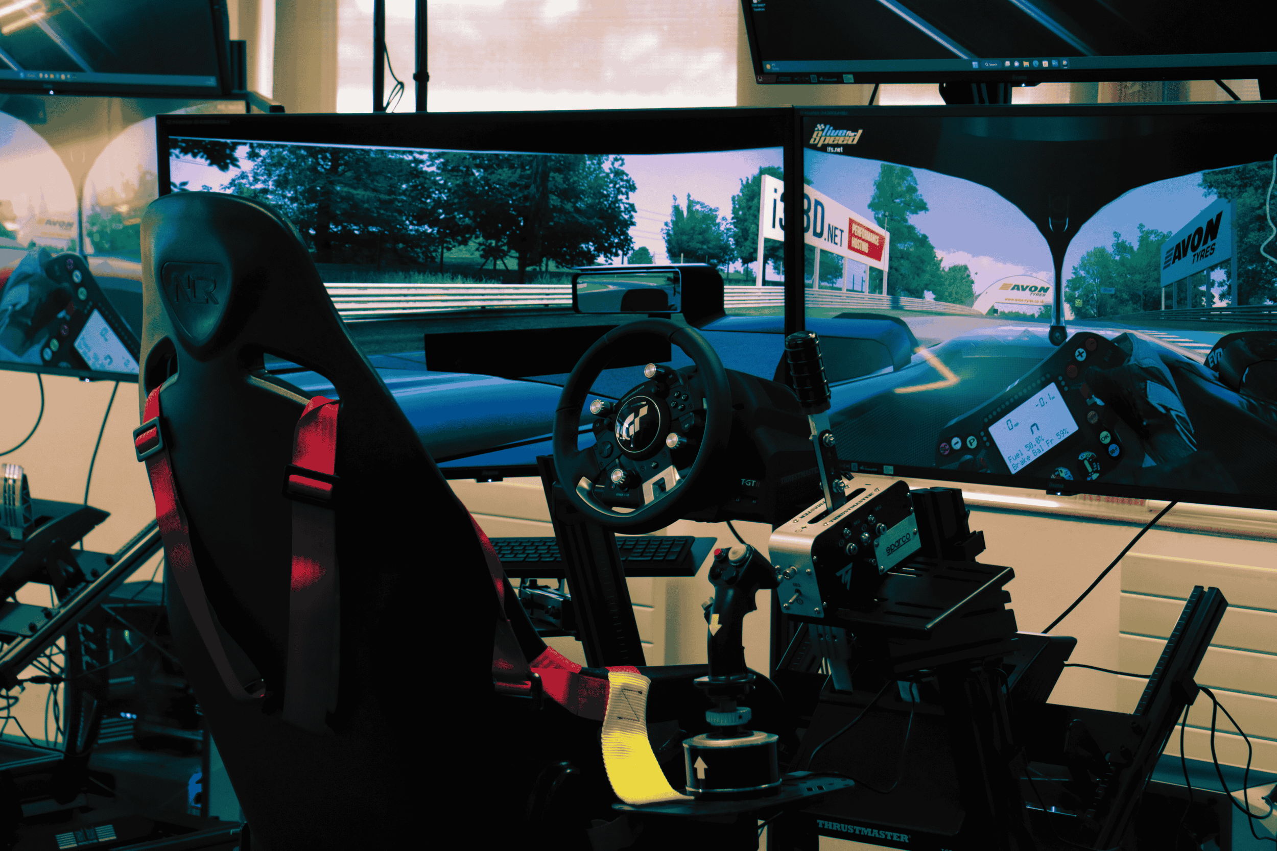 Racing simulation setup with a racing seat, steering wheel, pedals, multiple monitors displaying a race track scene, and various control devices.