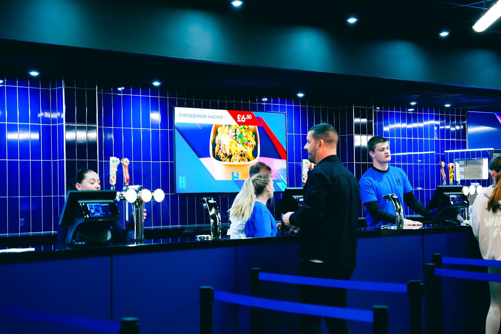 Inside a modern fast-food restaurant with blue tiled walls, two cashiers, and two customers at the counter. A digital menu displays a cheeseburger nachos for £6.50.