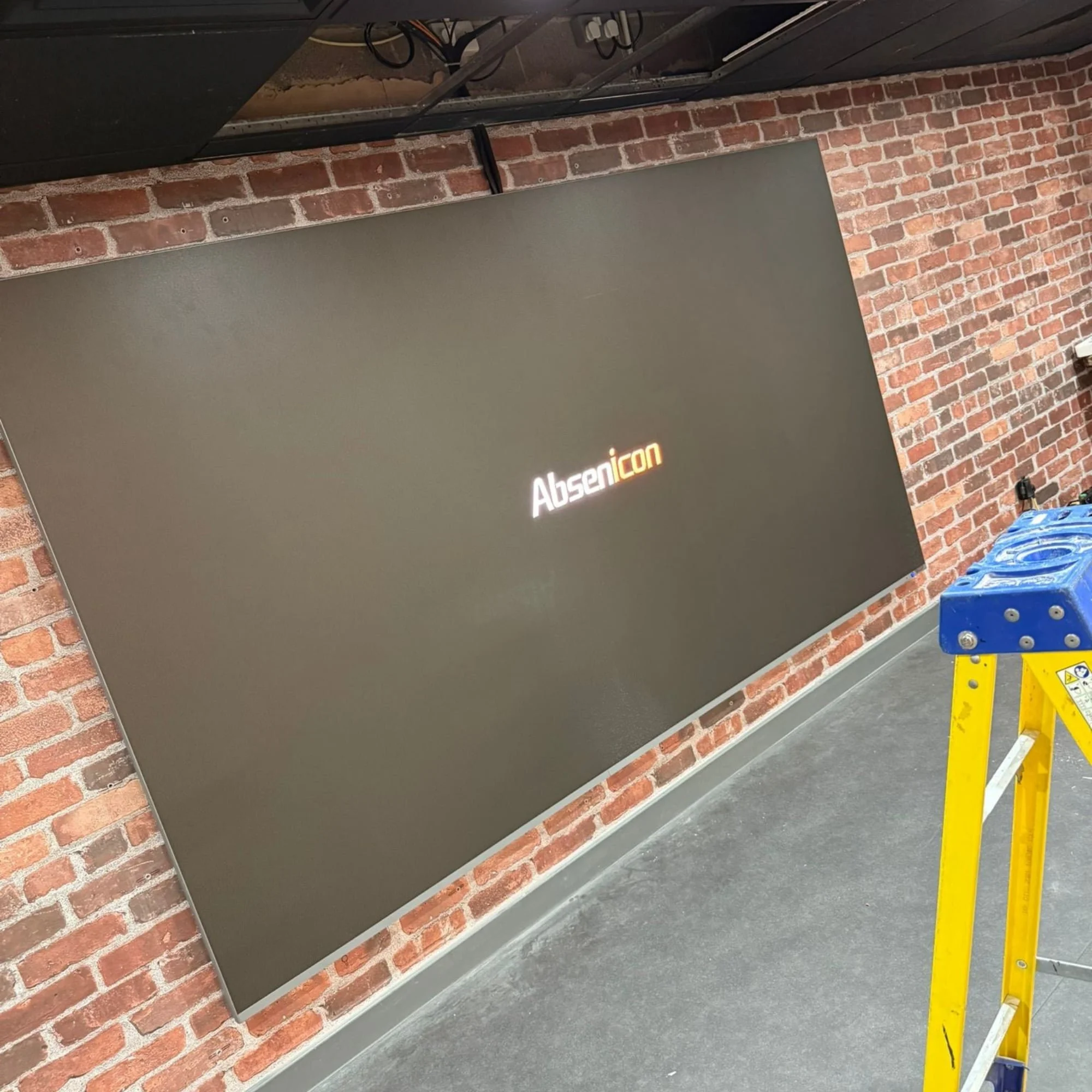 A large flat screen monitor displaying the logo 'AbsenIcon' mounted on an exposed brick wall in an industrial-style interior, with a yellow step ladder in the foreground.