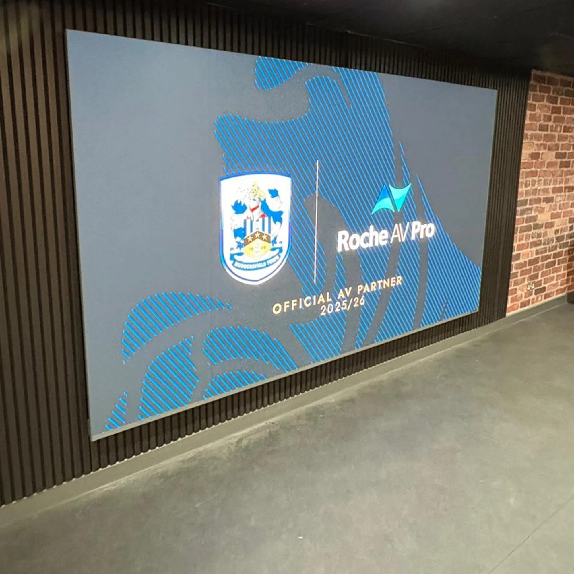 Digital sign on wall showing logos of a football team crest and Roche AV/Pro, with text indicating they are official AV partners for the 2025/26 season.
