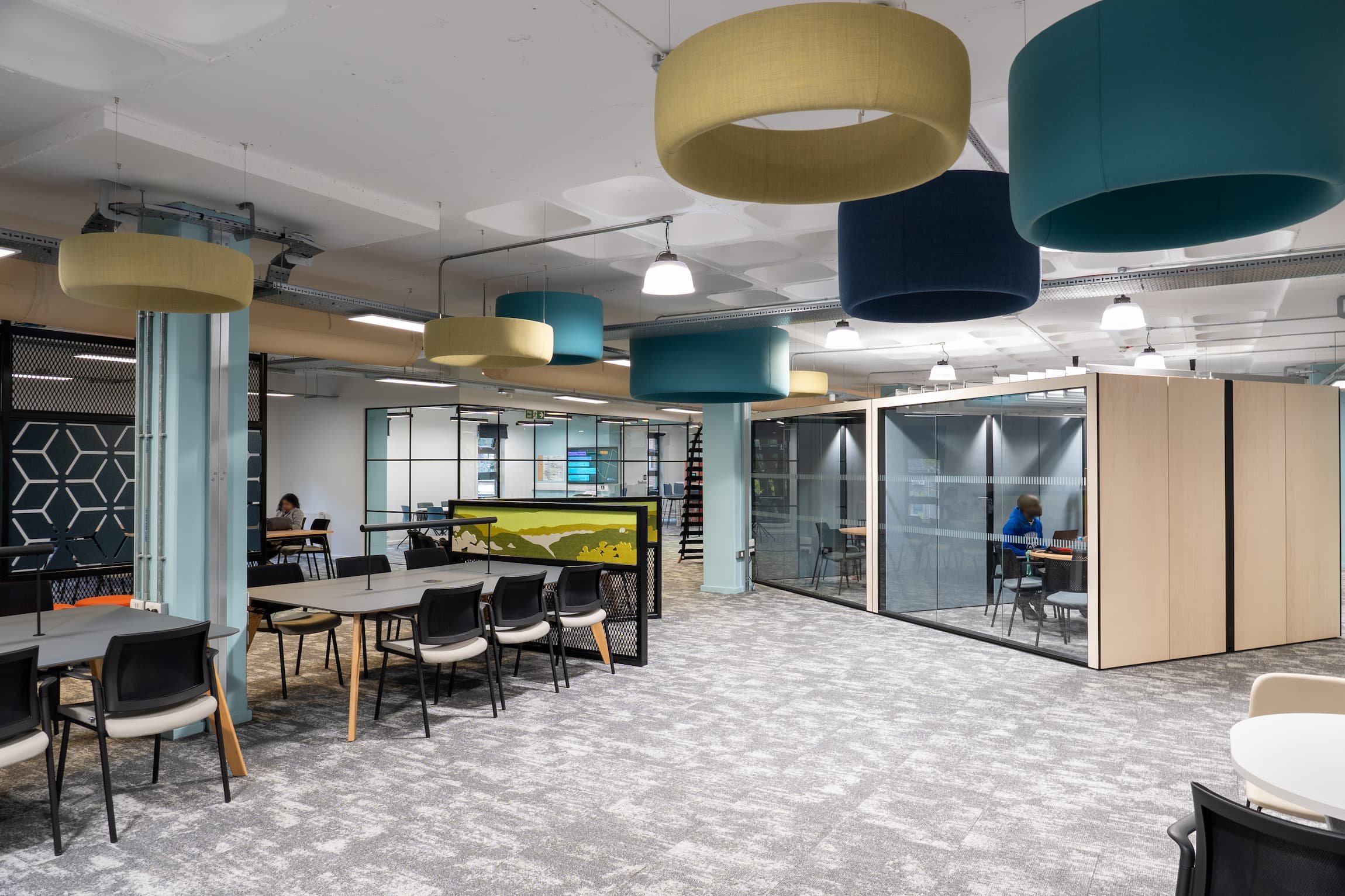 Modern open-plan office space with colorful hanging ceiling fixtures, glass-walled meeting rooms, and workstations with chairs.