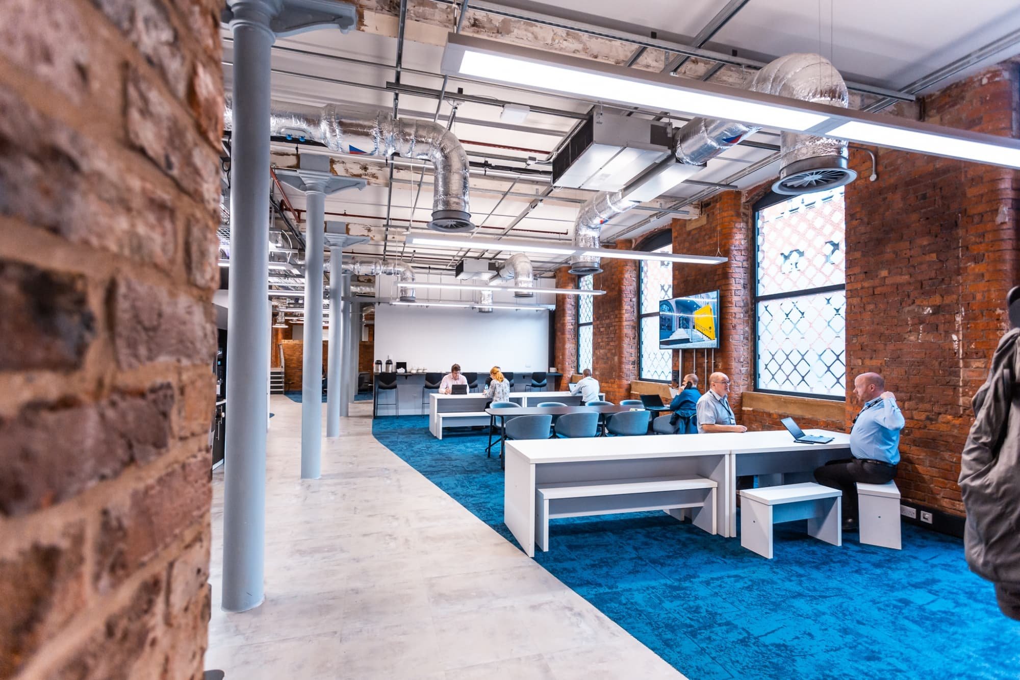 Modern office space with exposed brick walls, large windows, and work desks. Several people are working at their desks, and a large screen displays a presentation or video. The space has a blue carpet and industrial-style ceiling with visible ductwor