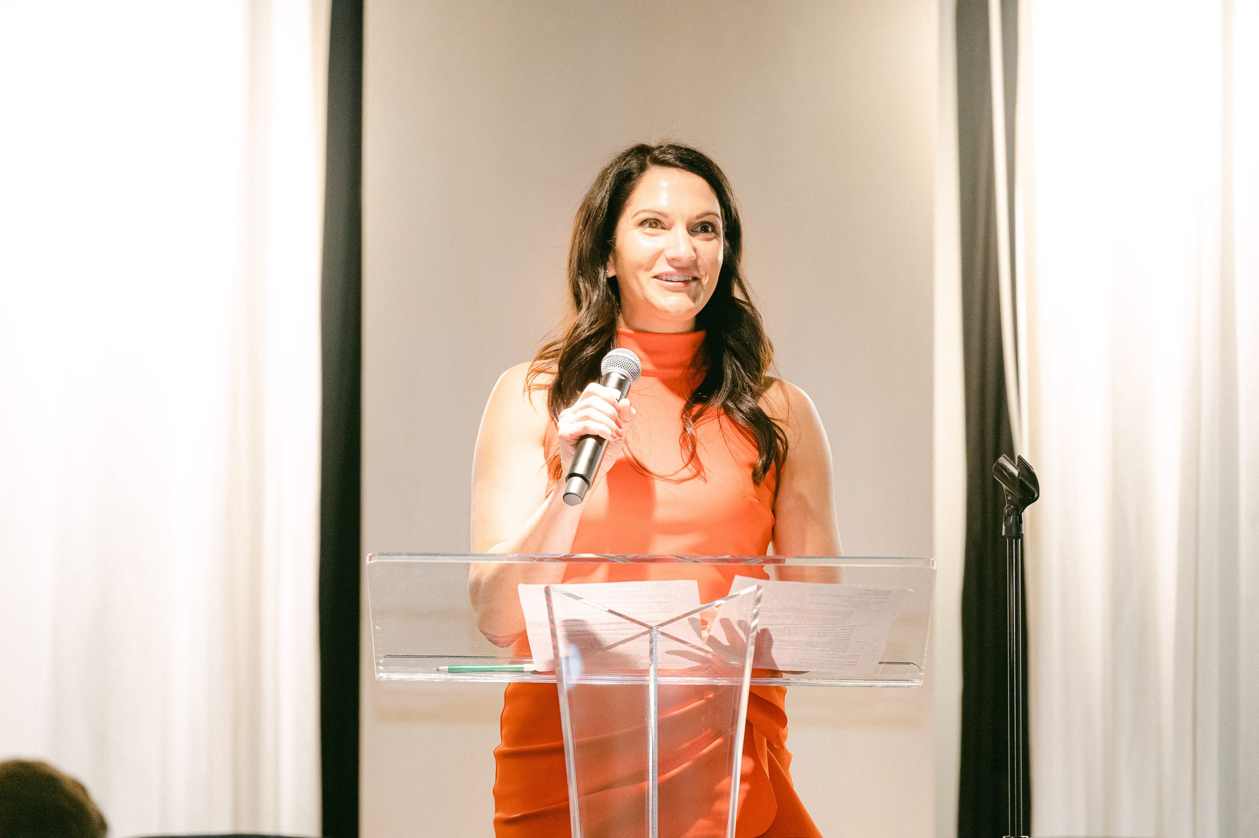 Woman in orange sleeveless dress speaking into a microphone at a podium.