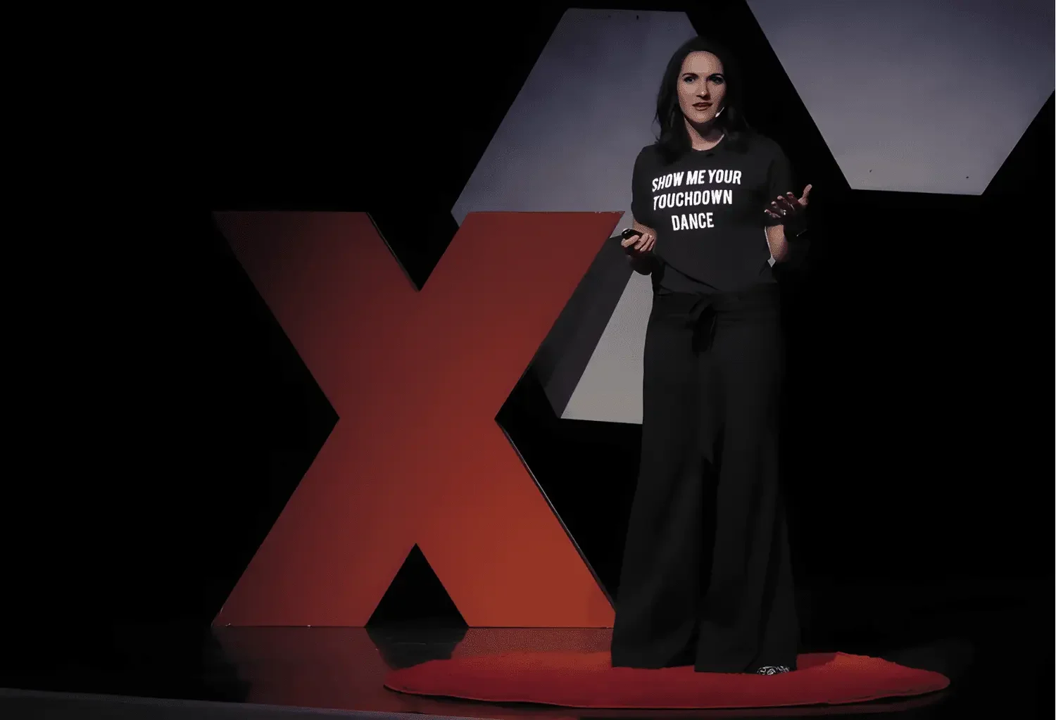 Amy Siegfried, TED speaker on stage at TEDx event, wearing a black T-shirt with white text, standing next to a large red 'X' and speaking to the audience.