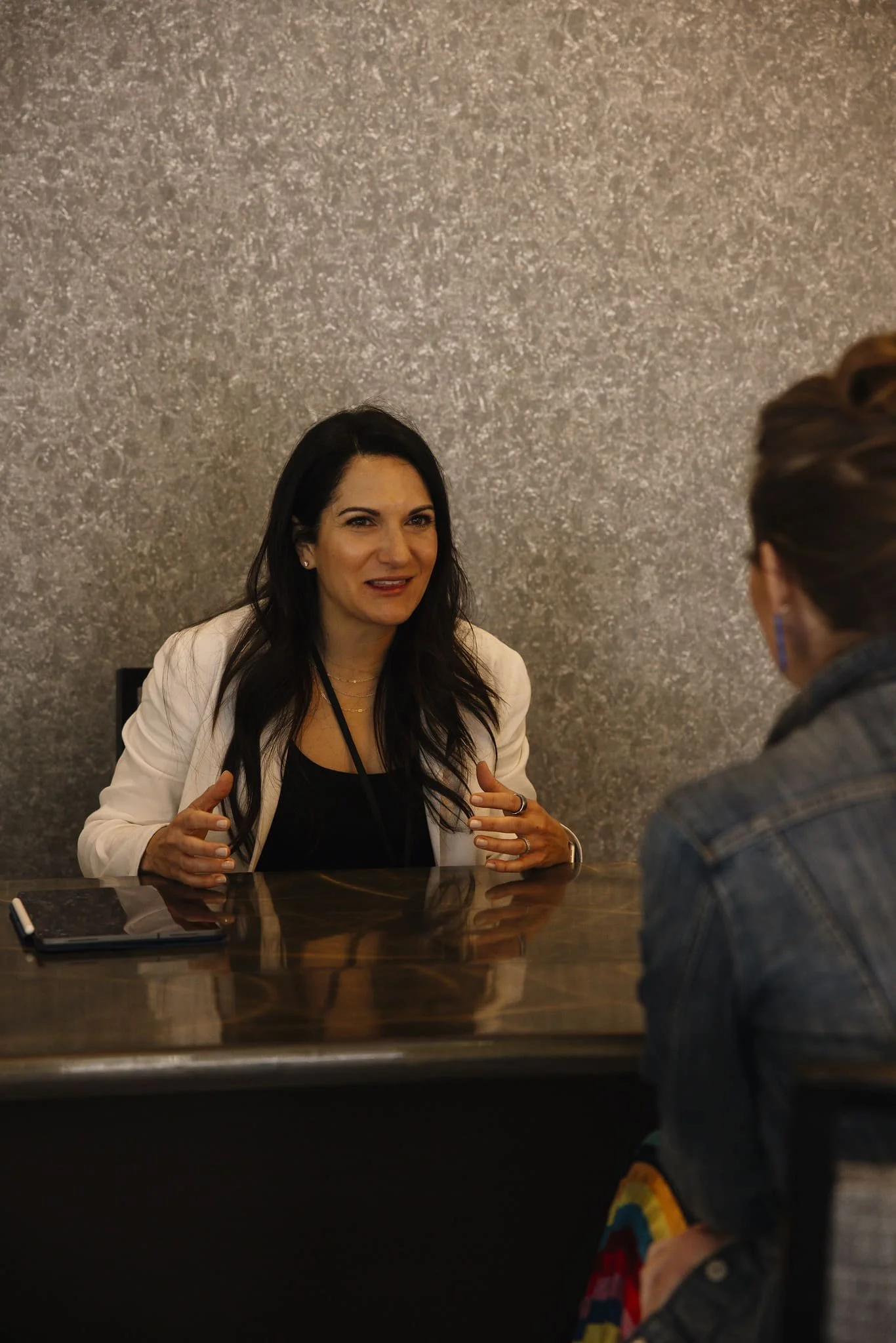 A woman with long dark hair, wearing a white blazer over a black top, is speaking and gesturing with her hands while seated at a polished wooden table. There is a tablet on the table and another person, wearing a denim jacket, is sitting across from her. The background is a textured gray wall.