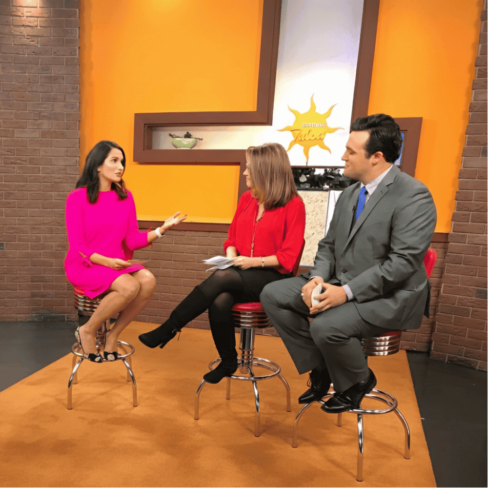 Three people in a TV studio: a woman in a pink dress, a woman in a red blouse, and a man in a gray suit sitting on stools, engaged in conversation during a talk show. Behind them is a yellow-orange wall with a sun graphic and TV screens.