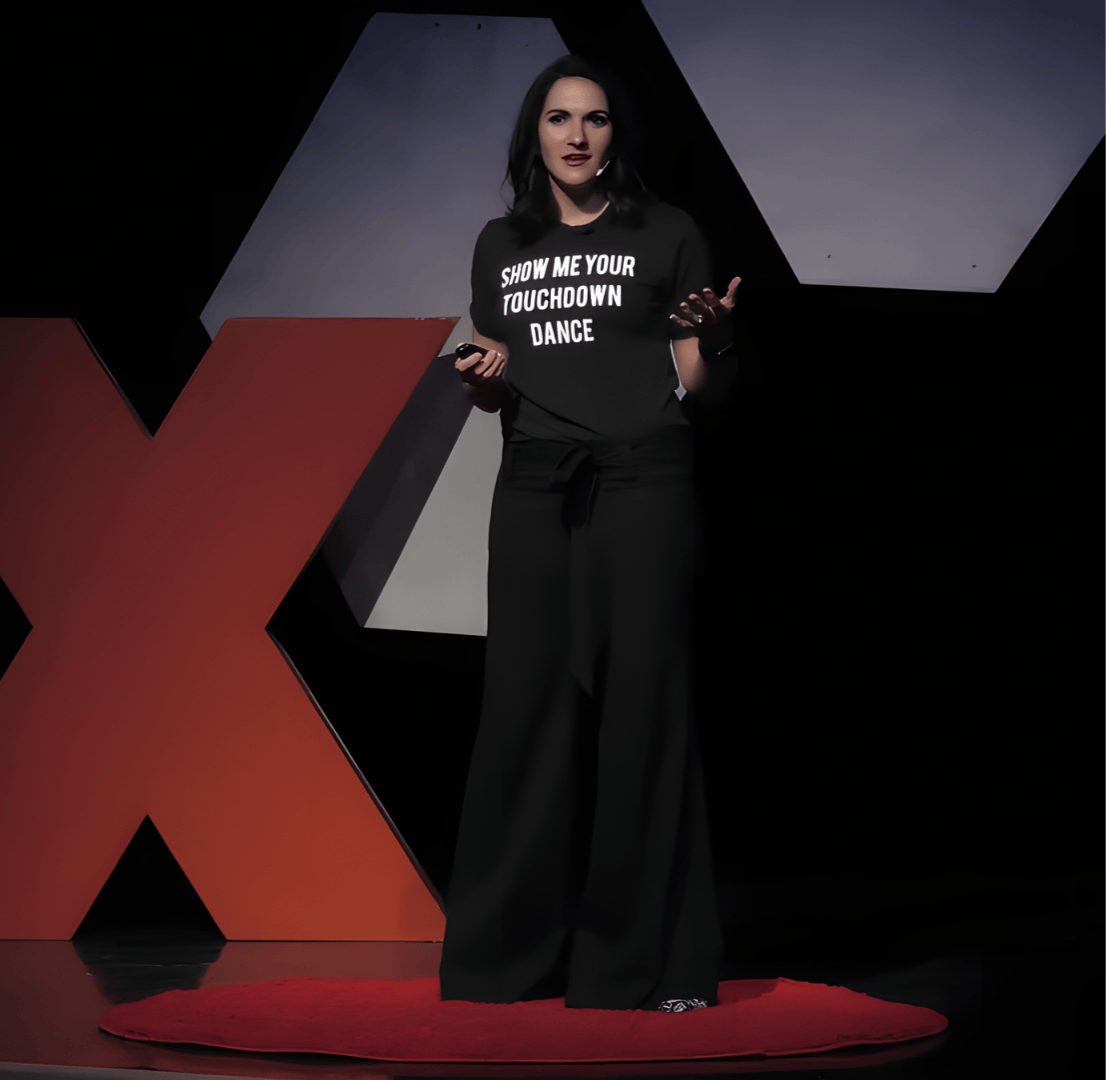 A woman standing on a red circular carpet, giving a presentation on a stage with large red and gray geometric shapes behind her. She is wearing a black T-shirt that says 'SHOW ME YOUR TOUCHDOWN DANCE' and black pants.