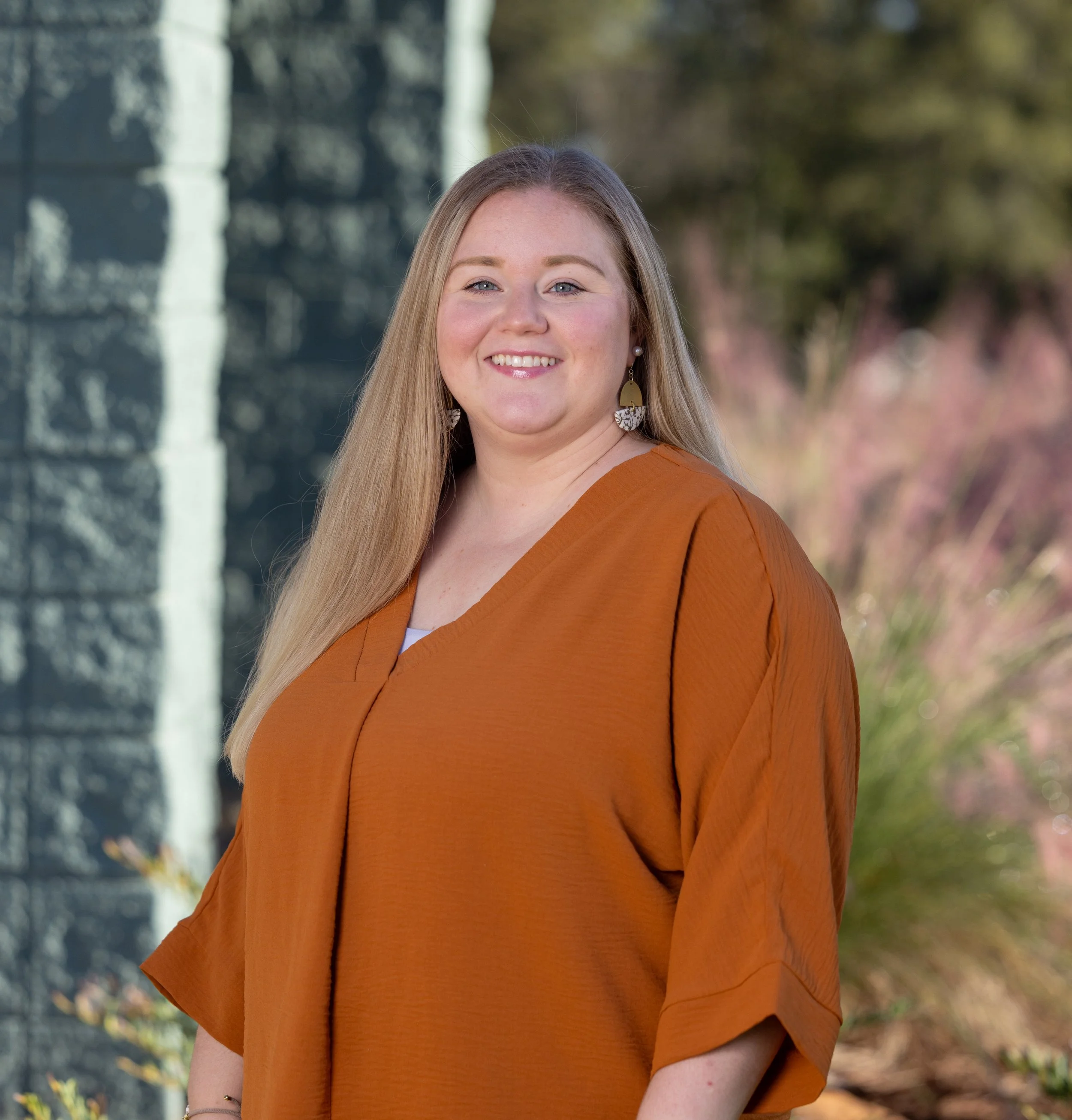 A woman smiling, standing outdoors against a blue-green wall, wearing a white top, turquoise earrings, and a name tag.