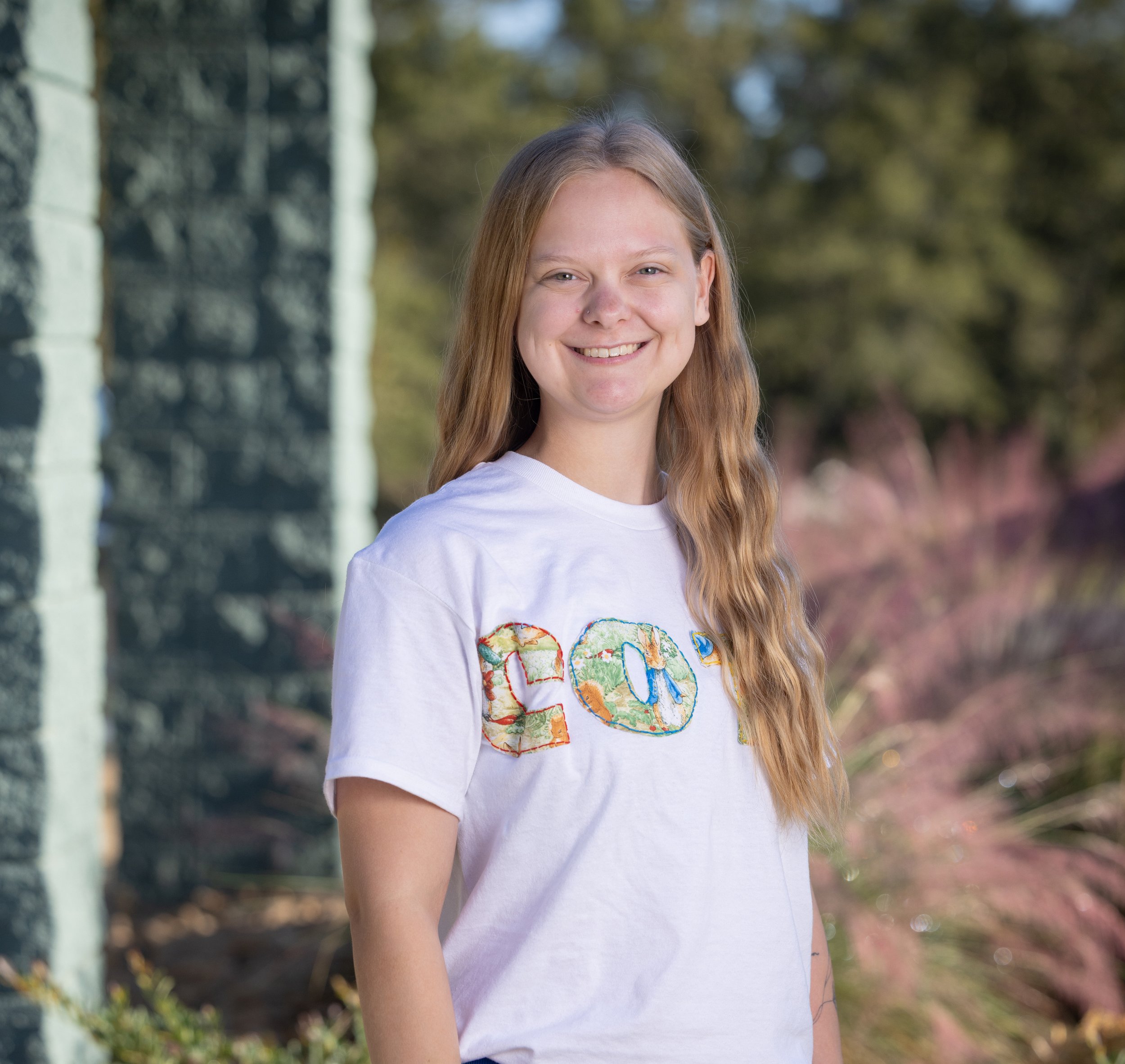 A young woman with long red hair, wearing a tan headband, black shirt, earrings, a necklace, and a tattoo on her right arm, smiling against a white background.