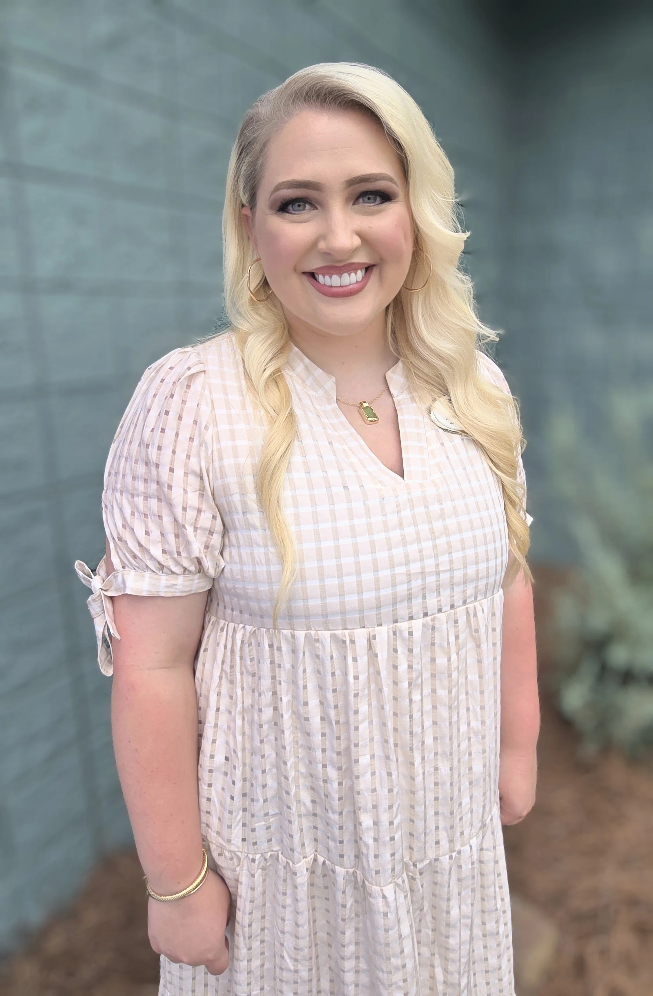 A woman with long blonde hair, wearing a beige and white checkered dress, smiling outdoors in front of a blue-green wall.