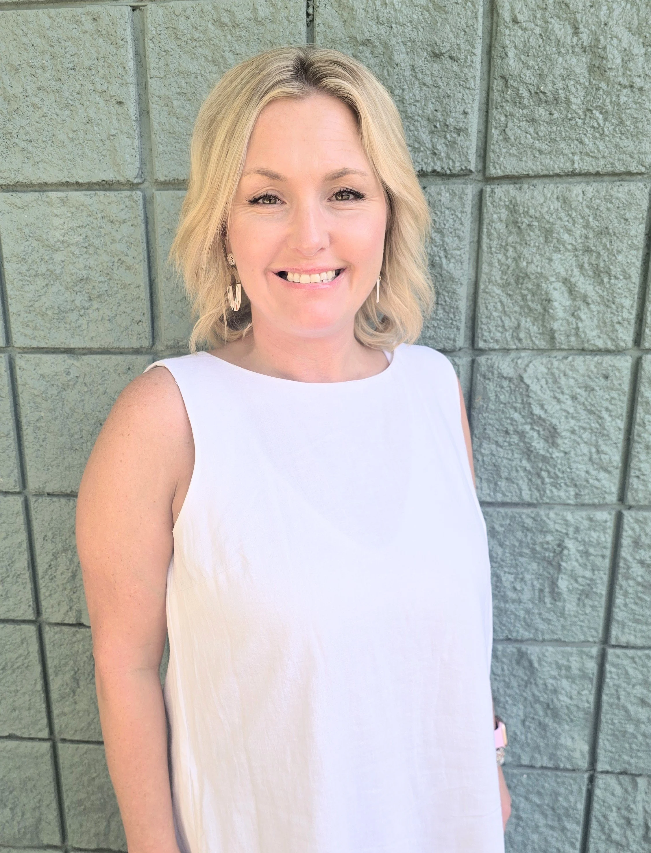 A woman with blonde hair, wearing a white sleeveless dress, smiling and standing against a green brick wall.