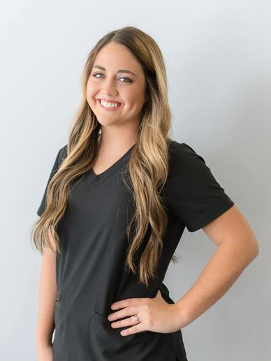 Smiling young woman with long blonde hair wearing black scrubs, standing with her hand on her hip against a light gray background.