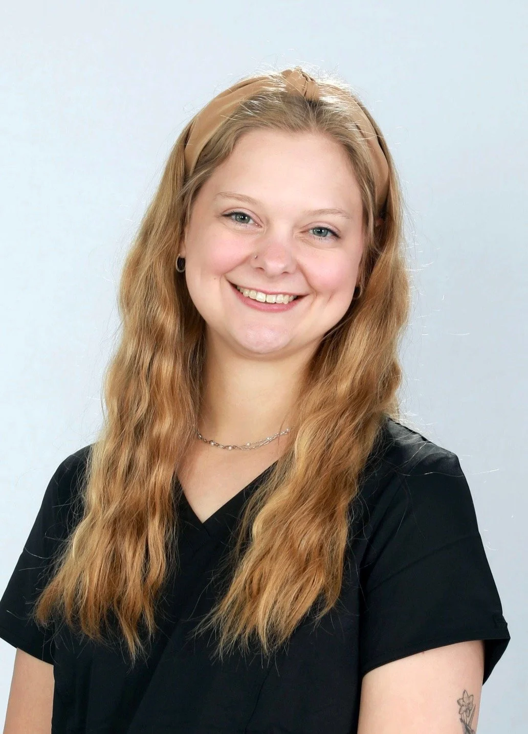 A young woman with long red hair, wearing a tan headband, black shirt, earrings, a necklace, and a tattoo on her right arm, smiling against a white background.