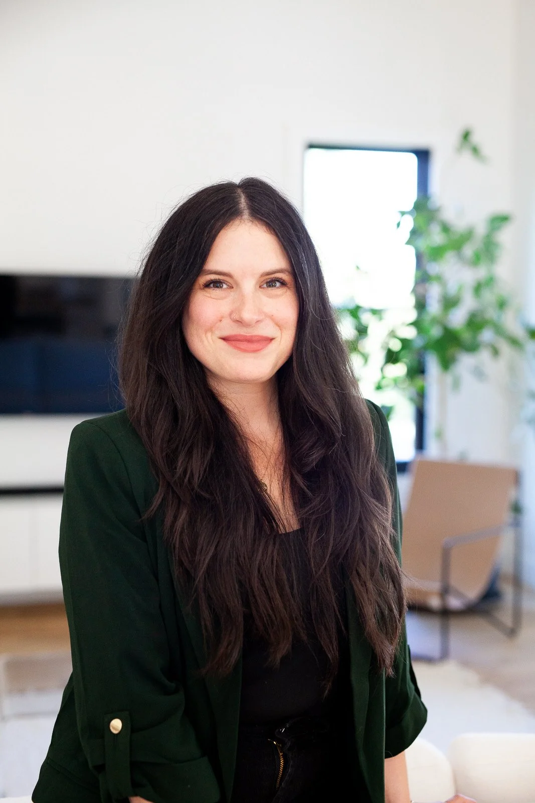 A woman with long dark hair smiling in a modern living room with plants and a beige chair in the background.