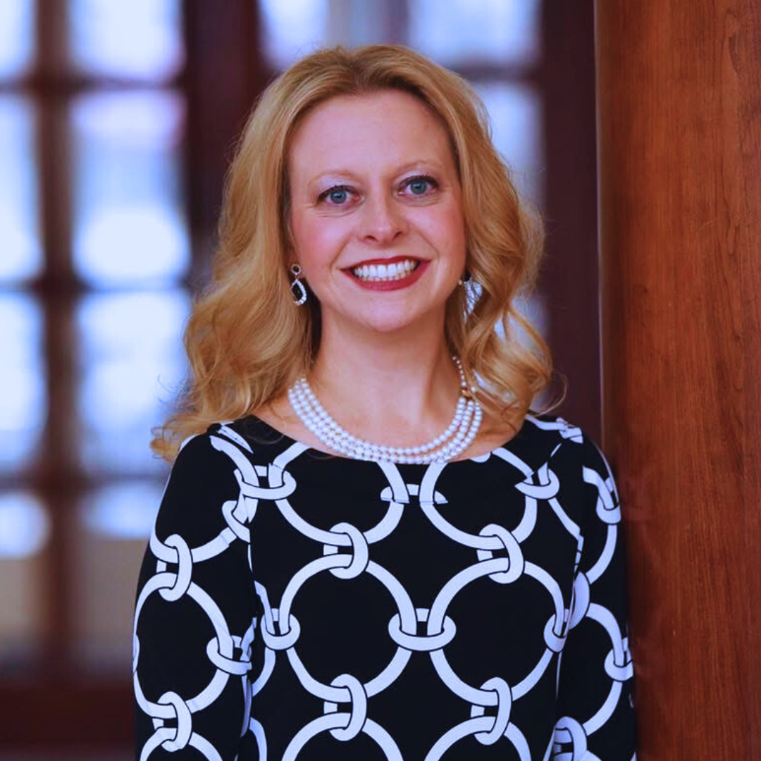 A woman with blonde hair smiling, wearing a black dress with white chain-link pattern, pearl necklace, and earrings, standing indoors near a wooden pillar.