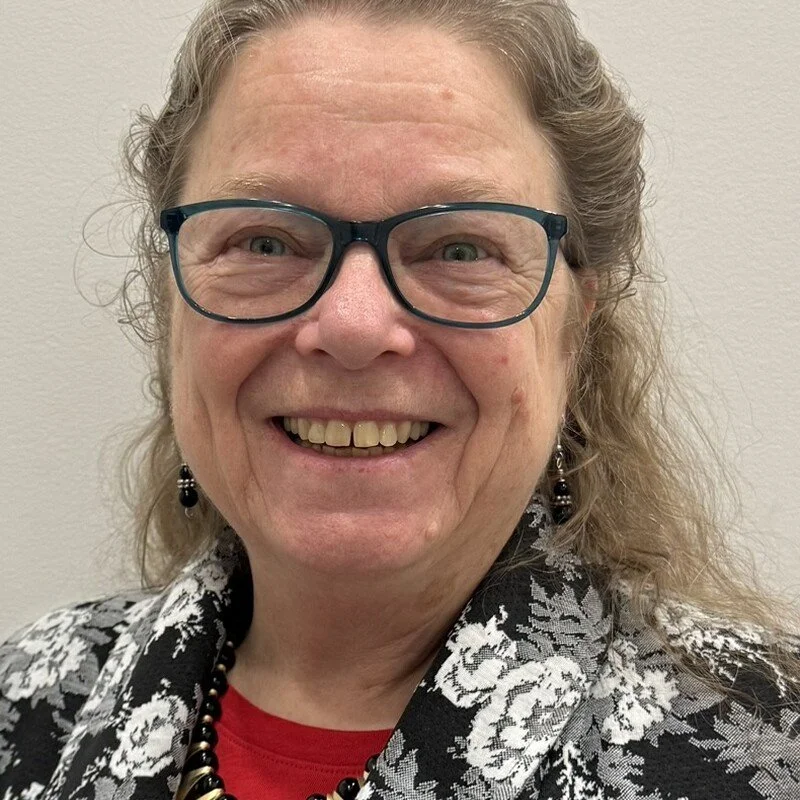 A smiling woman with long curly hair, wearing glasses, earrings, a floral patterned jacket, and a red shirt, against a plain white background.