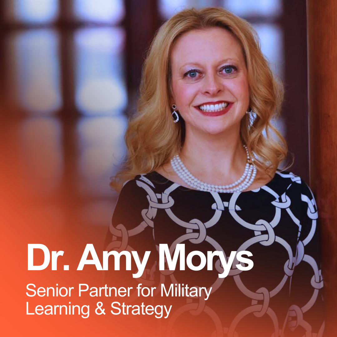 A woman with blonde hair smiling, wearing a black and white patterned dress, pearl necklace, and earrings, standing indoors with a blurred background. Text overlay reads 'Dr. Amy Morys, Senior Partner for Military Learning & Strategy.'