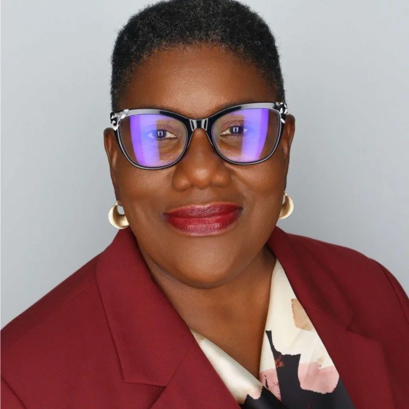 A woman with short curly hair wearing glasses with blue reflection, red lipstick, pearl earrings, a maroon blazer, and a beige blouse with a pattern, smiling against a light gray background.