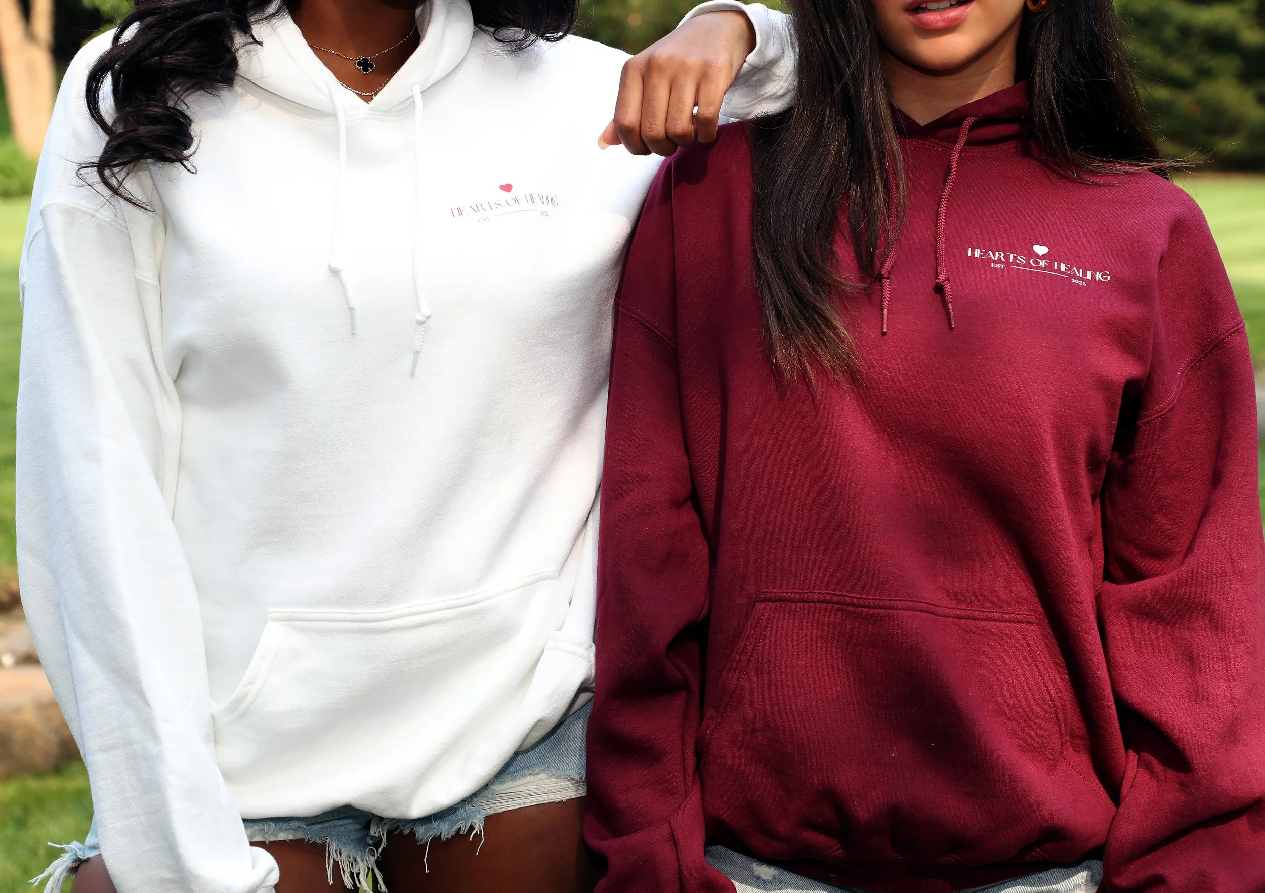 Two women wearing hoodies with the words 'Hearts of Healing' on them, standing outdoors in a park or garden.
