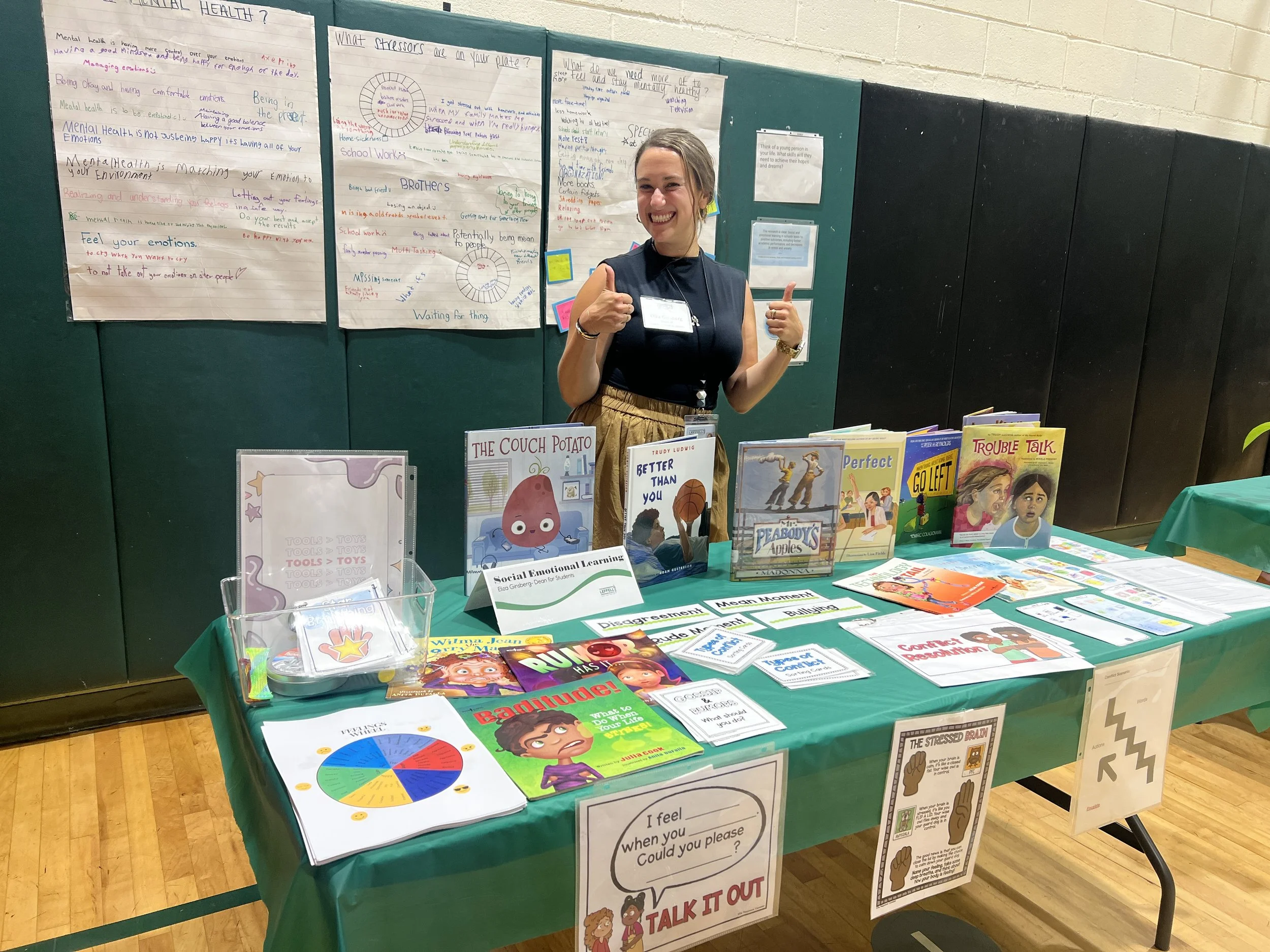 A woman at an educational booth featuring children's books and displays on social-emotional learning. She is smiling and giving a thumbs-up. The booth is set up in a gymnasium or classroom with posters about mental health and emotional wellness behind her.